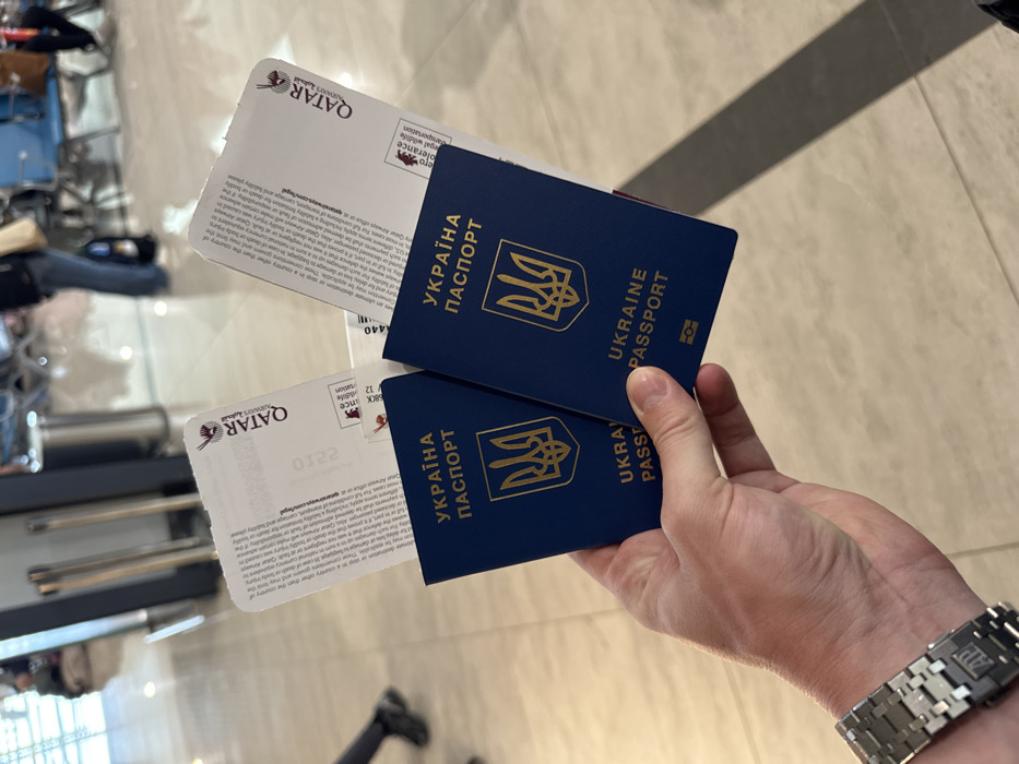 A close-up image depicts a hand, identified as male by its visible wrist and forearm, holding two dark blue Ukrainian passports. Each passport displays the gold Ukrainian Trident emblem and the text "УКРАЇНА ПАСПОРТ". Partially visible behind each passport are white Qatar Airways boarding passes, bearing the airline's logo and small print. The hand wears a metallic wristwatch with a geometric link bracelet. The background, blurred, reveals an indoor airport terminal environment with a light-colored tiled floor, chrome stanchions, blue airport-style seating, and several indistinct individuals, some seated and one walking. This scene is located within an airport terminal in Zanzibar, Tanzania.