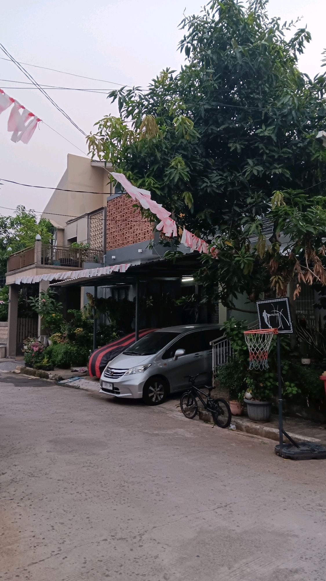The image depicts a residential street in Depok, Indonesia, under an overcast sky. A silver minivan is parked on the paved road in front of a two-story house. The house features a dark grey roof extending over a carport, with decorative red brickwork visible on an upper balcony. Pink and white pennant flags are strung across the front of the house, draped over wires and tree branches. To the right of the minivan, a black bicycle is parked on the side of the road. A basketball hoop with a red net is set up on the property, near the house and a lush green tree. The foreground shows a textured concrete road surface.
