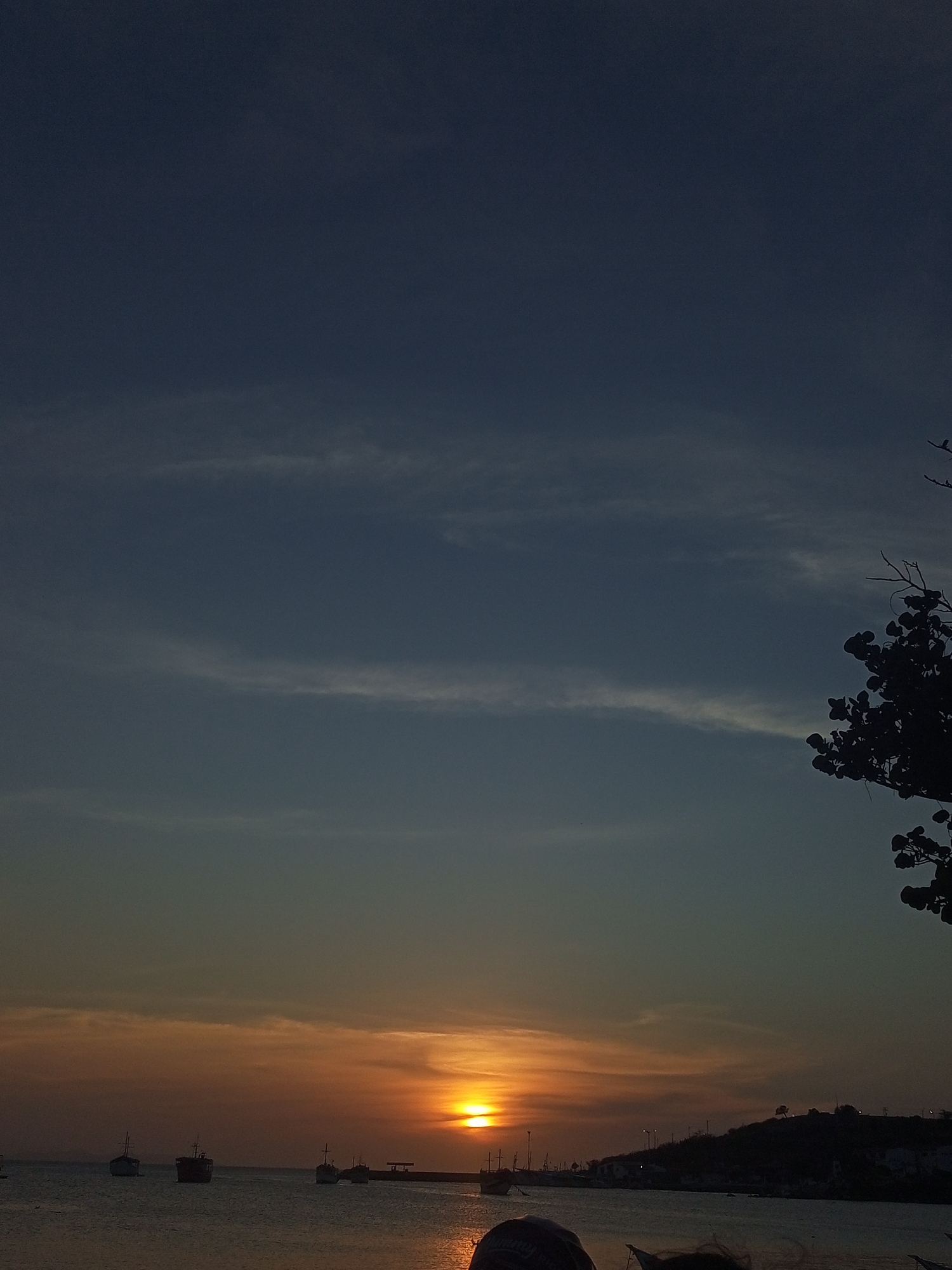 The image captures a sunset in Juan Griego, Venezuela. The sun is setting over the water, casting an orange glow across the horizon. Several boats are silhouetted against the water. In the distance, the outline of land with some buildings and streetlights can be seen. In the right upper quadrant of the image there are silhouetted tree branches. The sky transitions from a darker blue above to a lighter hue closer to the horizon.