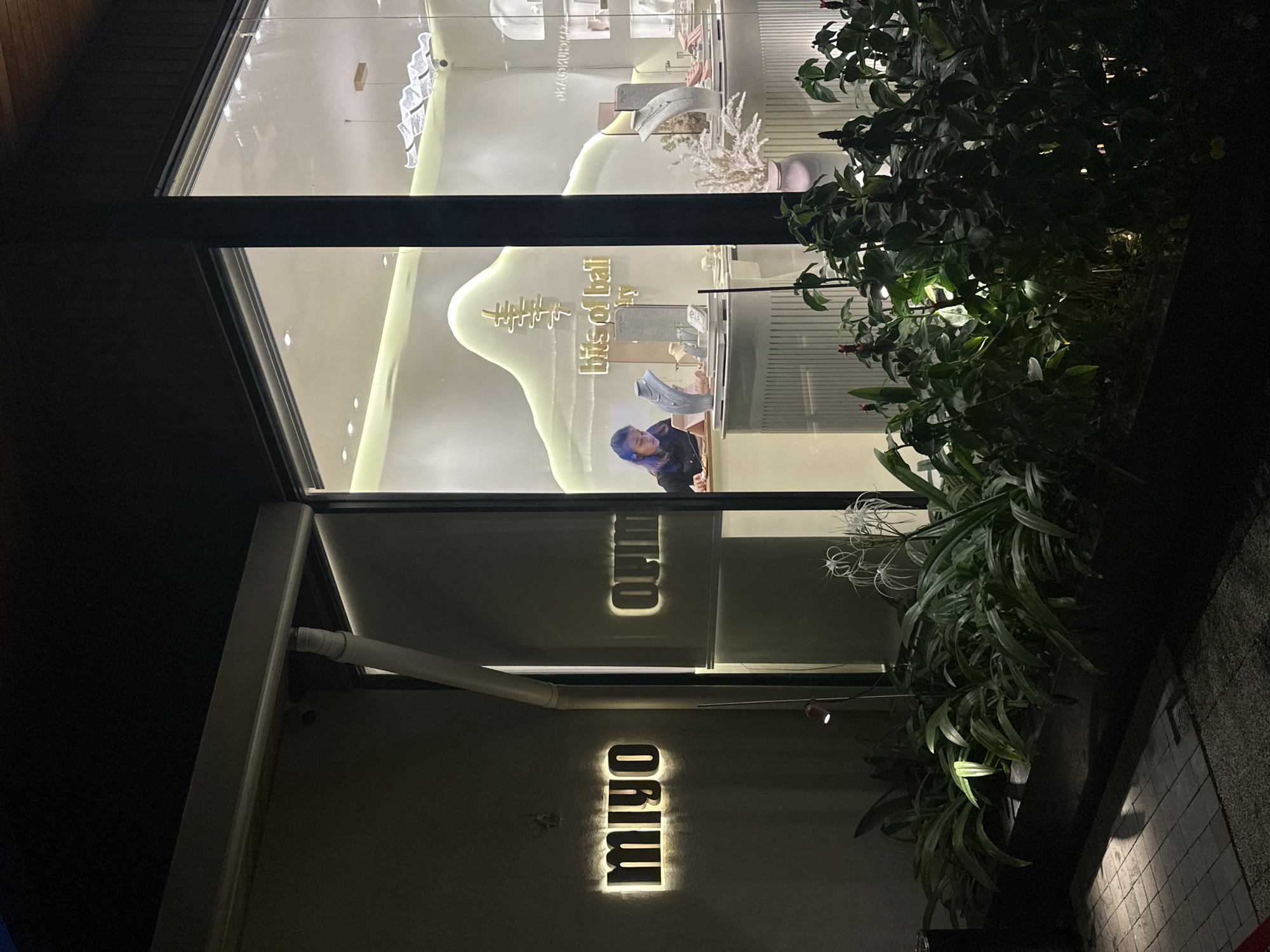This is a nighttime, exterior shot of a modern shop or boutique. The image is framed vertically, showcasing a large, brightly lit display window.
Inside the shop, a person with purple hair, wearing a dark top, is seen leaning over a counter or display case, seemingly engaged with items. The interior display features various small objects, possibly jewelry or decorative pieces, arranged on white, undulating stands. Above these displays, illuminated text reads "bits of ball," alongside a stylized graphic resembling a tree or coral. The shop's interior is brightly lit, contrasting with the dark exterior.
To the right of the building, lush, green foliage, including plants with red buds and white, spidery flowers, grows in planter boxes, appearing well-maintained. On the building's facade, below the main display window, an illuminated sign spells out "migo." A clear, inverted reflection of this sign is visible on the glass directly above it. Dark architectural beams frame the windows and the overall structure. A tiled sidewalk is partially visible on the ground to the far right. The scene depicts a quiet evening at a stylish commercial establishment.