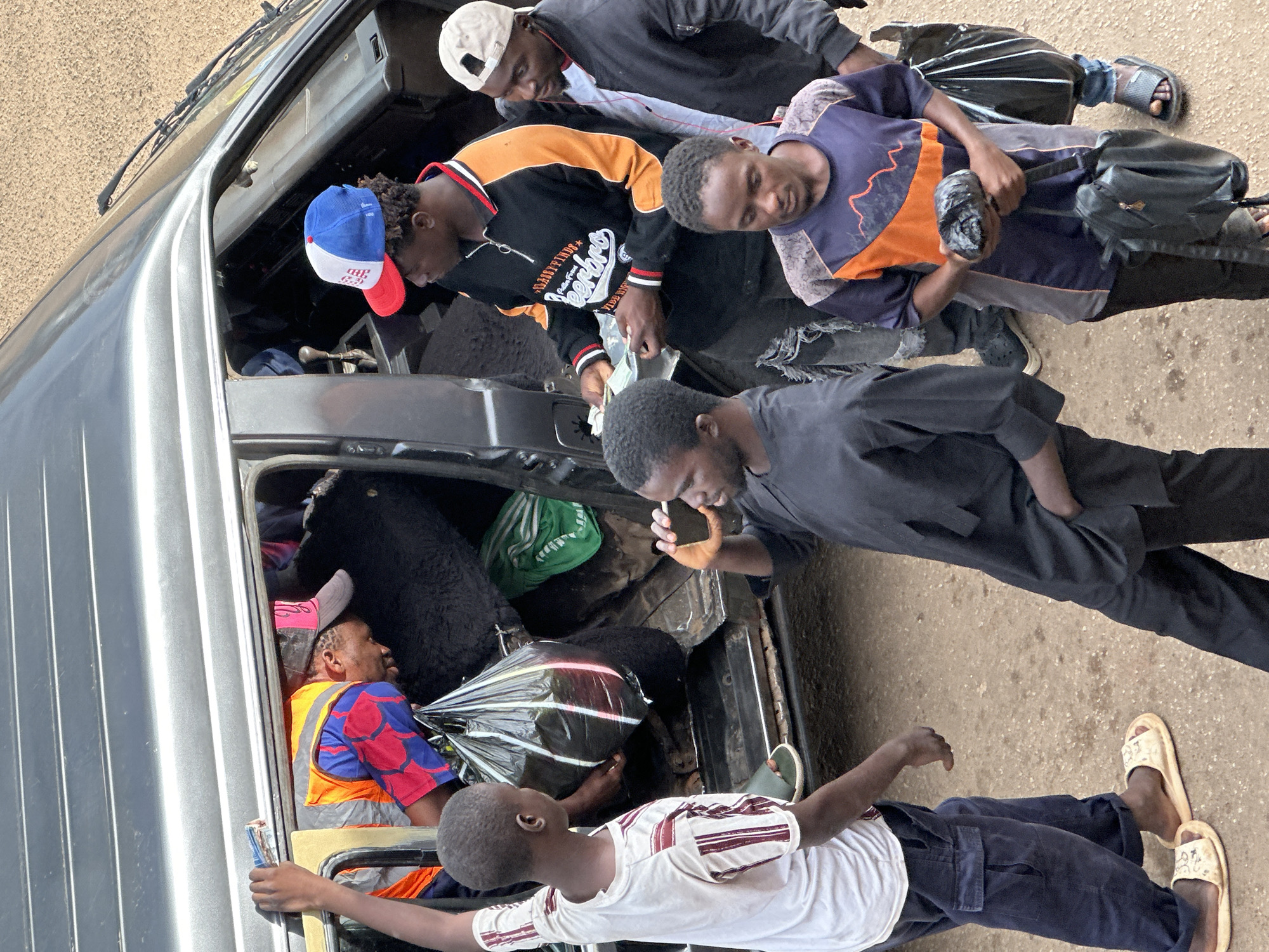 The image depicts a busy street scene in Bukuru, Nigeria, with several individuals engaged in various activities around a parked vehicle, possibly a van or a minibus. The vehicle is dark gray and appears to be open, with people loading or unloading items from its interior and the back. One person, wearing a bright orange safety vest over a blue and red patterned shirt, is leaning into the vehicle, seemingly assisting with the process. They are wearing a pink baseball cap. Another young person, wearing a white t-shirt with red stripes and dark blue trousers, stands to the left, holding a hand up and looking towards the vehicle.
In the foreground, a young man in a black outfit is looking at his phone. To his right, a man wearing a black long-sleeved shirt with orange and white accents and a red, white, and blue baseball cap is holding a wad of cash in his hands. He appears to be engaged in a transaction or is about to hand over the money. Another individual, wearing a black jacket and a white baseball cap, stands to the right of the man with the money, looking on. Further to the right, a young man in a blue and orange patterned shirt is holding a black plastic bag and a black bag on his shoulder. The ground is a dusty, sandy surface, typical of a street in many Nigerian cities. The overall impression is one of everyday commerce and activity.