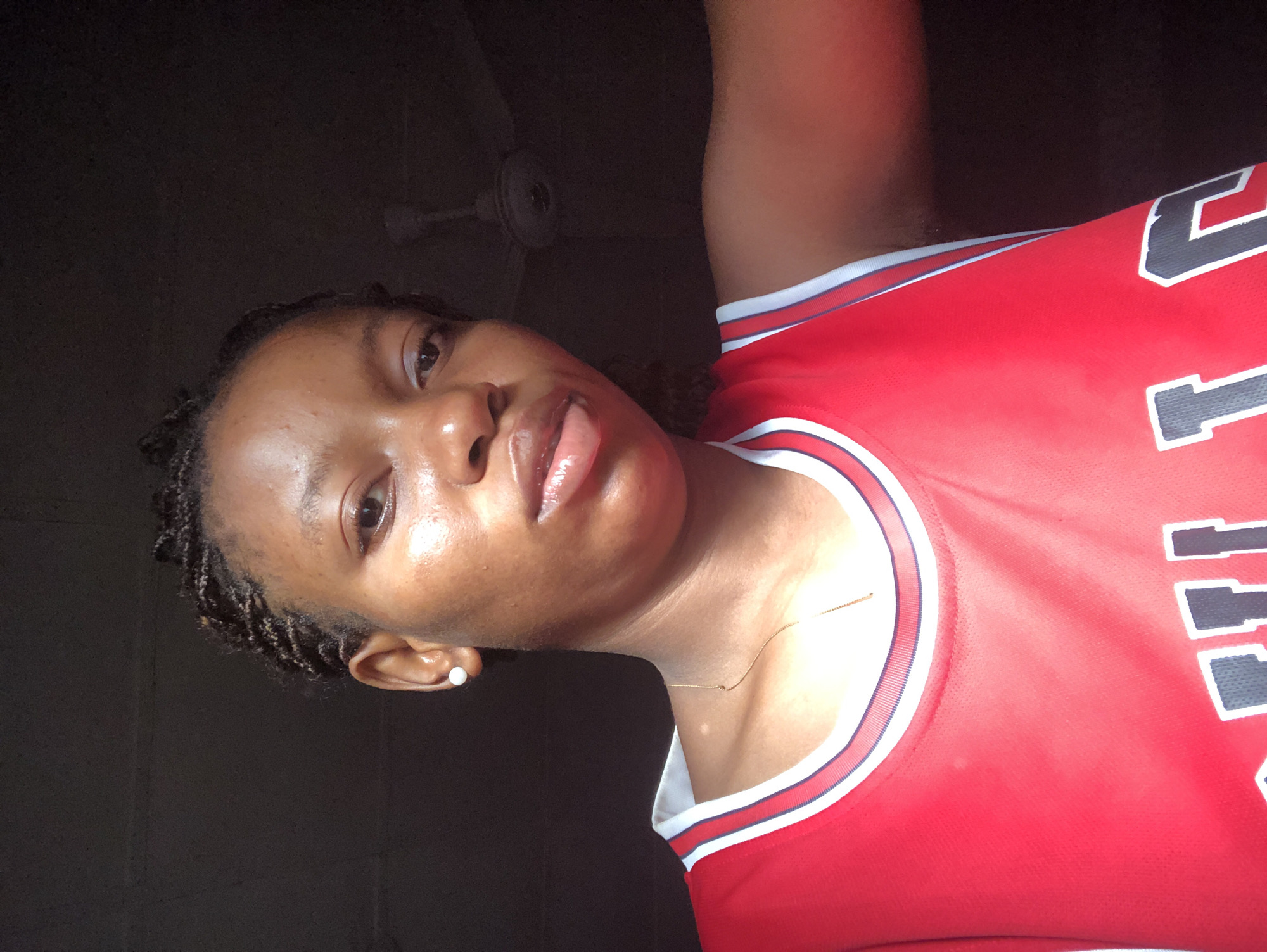 A dark-skinned person with braided hair is centered in the frame, facing the camera, wearing a red and white sports jersey with black lettering. They are indoors in a dimly lit room in Pindiga, Nigeria, with a ceiling fan visible in the background. The person's skin appears shiny, and they have a gold necklace and a single pearl earring.