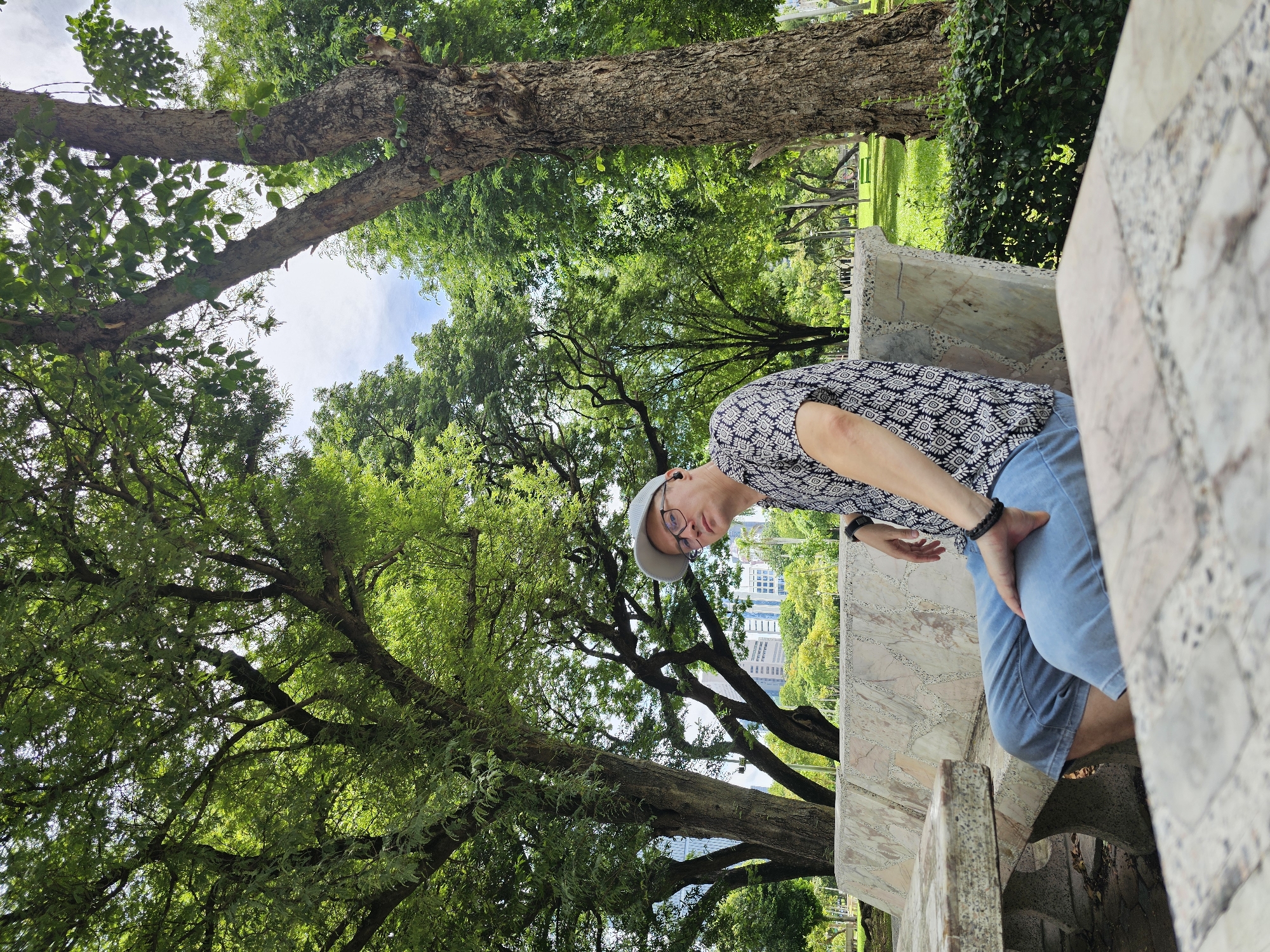 A man sits on a marble bench in a park in Bangkok, Thailand. He is wearing a light-colored cap, glasses, a patterned short-sleeved shirt, and blue jeans. He has on a black wristband and a black earbud in his left ear. Behind him, large trees with green foliage create a canopy. In the distance, modern buildings can be seen peeking through the trees. The park appears well-maintained with green grass and manicured bushes. The overall scene suggests a relaxed outdoor setting within the city.