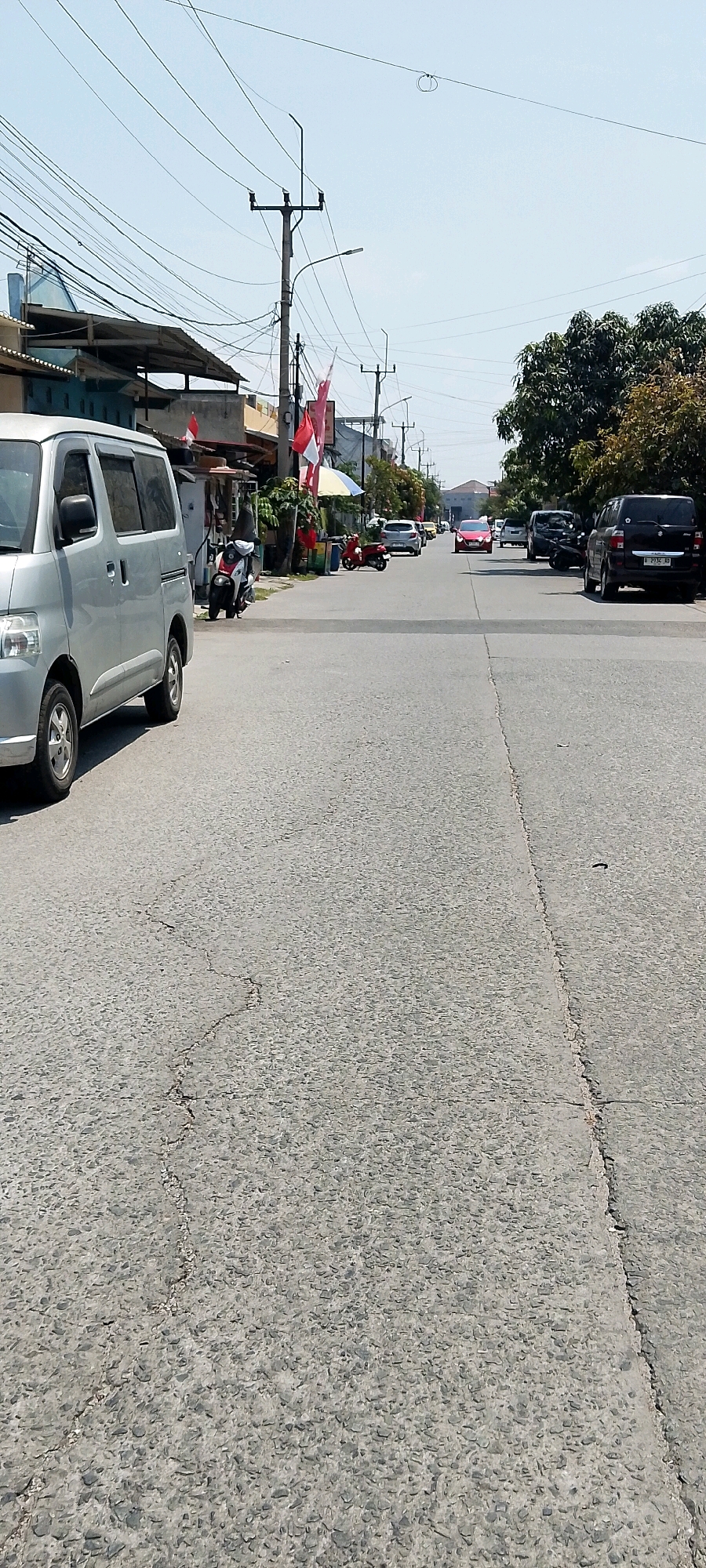 The image depicts a street scene in Satriamekar, Indonesia. The perspective is from a low angle, looking down a relatively wide asphalt road. The road surface is cracked and appears weathered. On the left side of the road, a silver minivan is parked, taking up a significant portion of the foreground. Behind the minivan, there are low-lying buildings with awnings and what appear to be storefronts. Several red and white Indonesian flags are visible, suggesting a patriotic or festive occasion. A few motorcycles are parked on the side of the road.
The road itself has a visible center line, and in the distance, several cars are traveling in the same direction. These include a red sedan in the middle of the road, followed by a dark SUV on the right side. Further down the road, more vehicles are present, including another SUV and a white car. Utility poles with numerous wires are present along the left side of the street, characteristic of urban infrastructure. Trees with dense green foliage are visible on the right side of the road, providing a contrast to the built environment. The sky is clear and bright blue, indicating a sunny day. The overall impression is that of a typical street in a developed area of Satriamekar, Indonesia, during daylight hours.