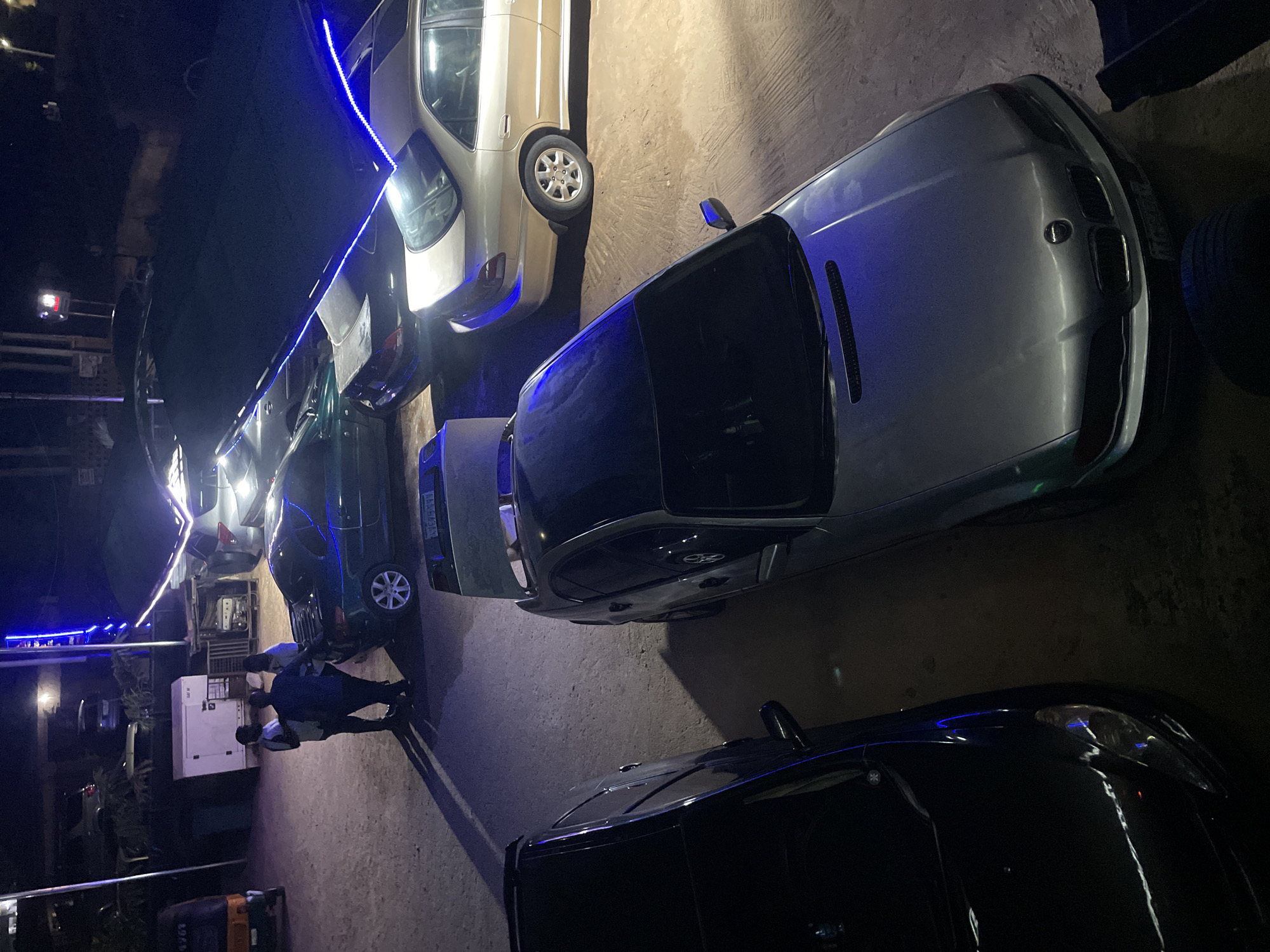 The image depicts a nighttime scene in Gura Top, Nigeria, showing multiple cars parked on a paved surface. The vehicles range in color from silver and gold to dark green and black. There are at least two people standing near a white generator. Blue neon lights run above the cars, illuminating the area. The setting appears to be a parking area or garage.