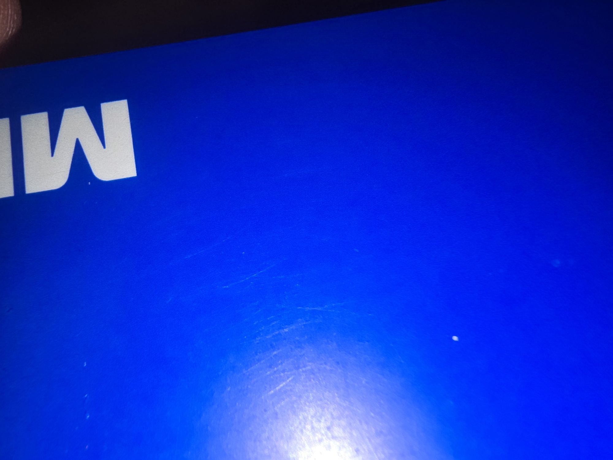 The image shows a close-up of a bright blue surface, likely a book cover or a similar object. The surface is marked with some faint scratches. Towards the top right corner, a portion of a white, sans-serif letter "M" is visible. The lighting suggests an indoor environment, with a bright light source on the left side of the image creating a glare and the right side of the image being darker. There are no discernible elements in the image that would allow for the determination of its location in Ulan-Ude, Russia, or any other specific geographical place.