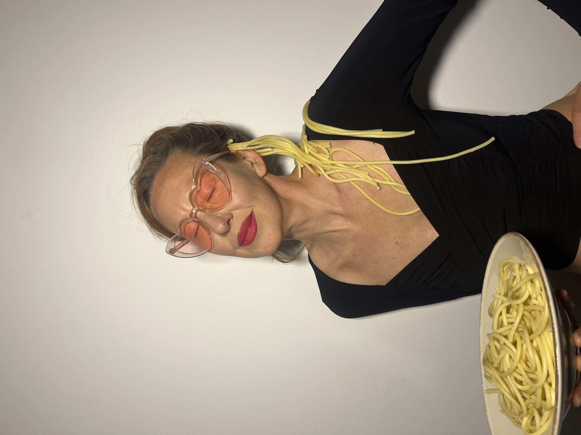 The image displays an adult female subject indoors against a plain, light-colored wall. The location is specified as the city of Budva, Montenegro. The subject has light brown hair pulled back, with some strands visible around her forehead. She is wearing large, translucent light pink heart-shaped sunglasses with pink lenses. Her eyes are closed, and she has red lipstick applied. She is dressed in a black, long-sleeved v-neck top with gathered fabric on the front. Cooked spaghetti strands are draped over her left ear, extending down her neck, across her left shoulder, and onto the front of her top. Her left hand holds a round, shallow, white ceramic plate with a light brown rim, filled with cooked spaghetti. Her right arm is bent, with the hand out of frame near her hip. The lighting is even and consistent, illuminating the subject and the background.