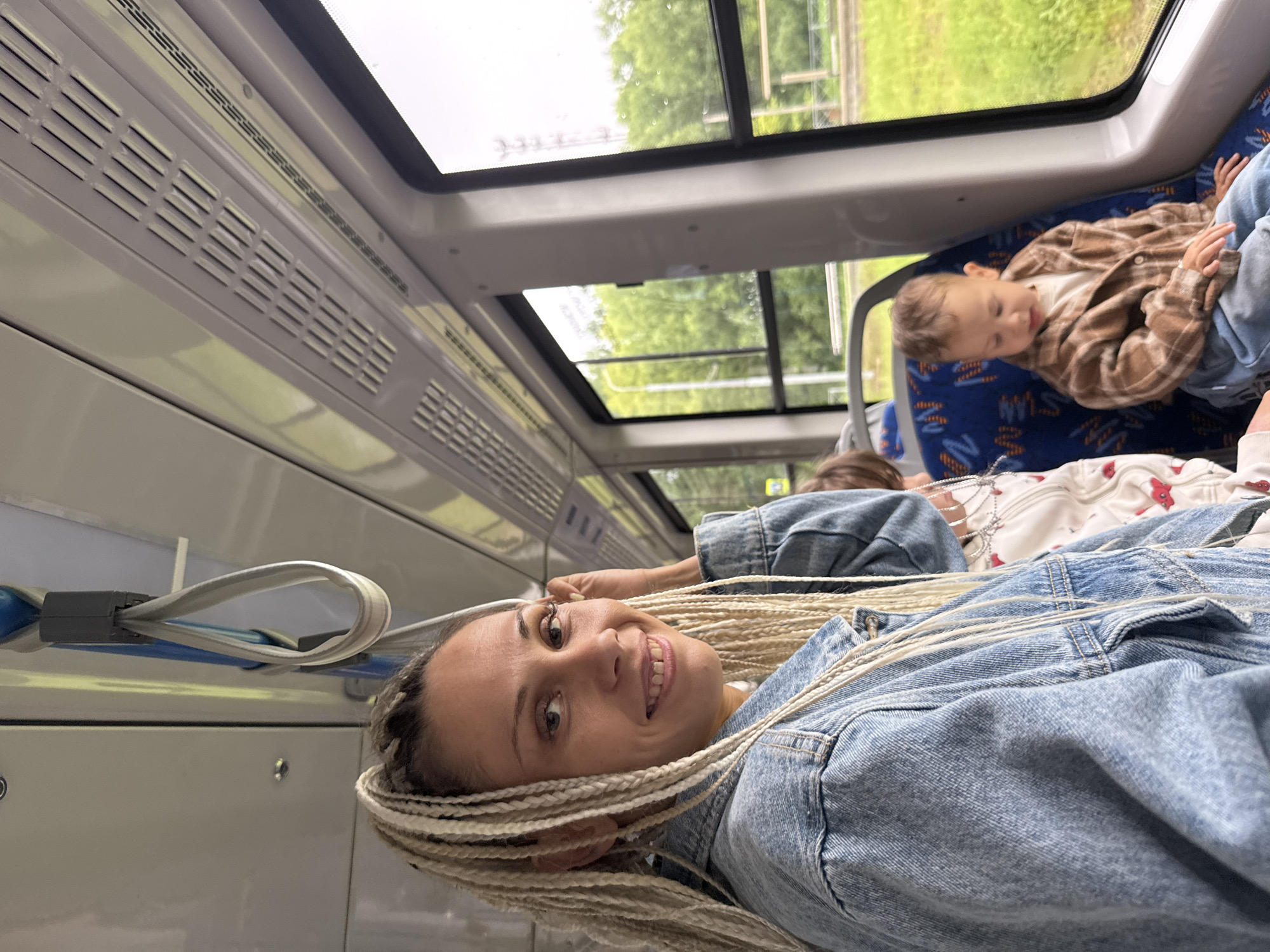 In Rusanovo, Russia, a woman with blonde braided hair, wearing a denim jacket, smiles at the camera while seated inside a train. She is in the foreground, with her head and shoulders visible. Behind her, a young child wearing a plaid shirt and blue jeans sits in a blue seat. The train's interior is visible, including overhead compartments, a window showing greenery outside, and seats. The woman is holding onto a strap attached to the train's ceiling.