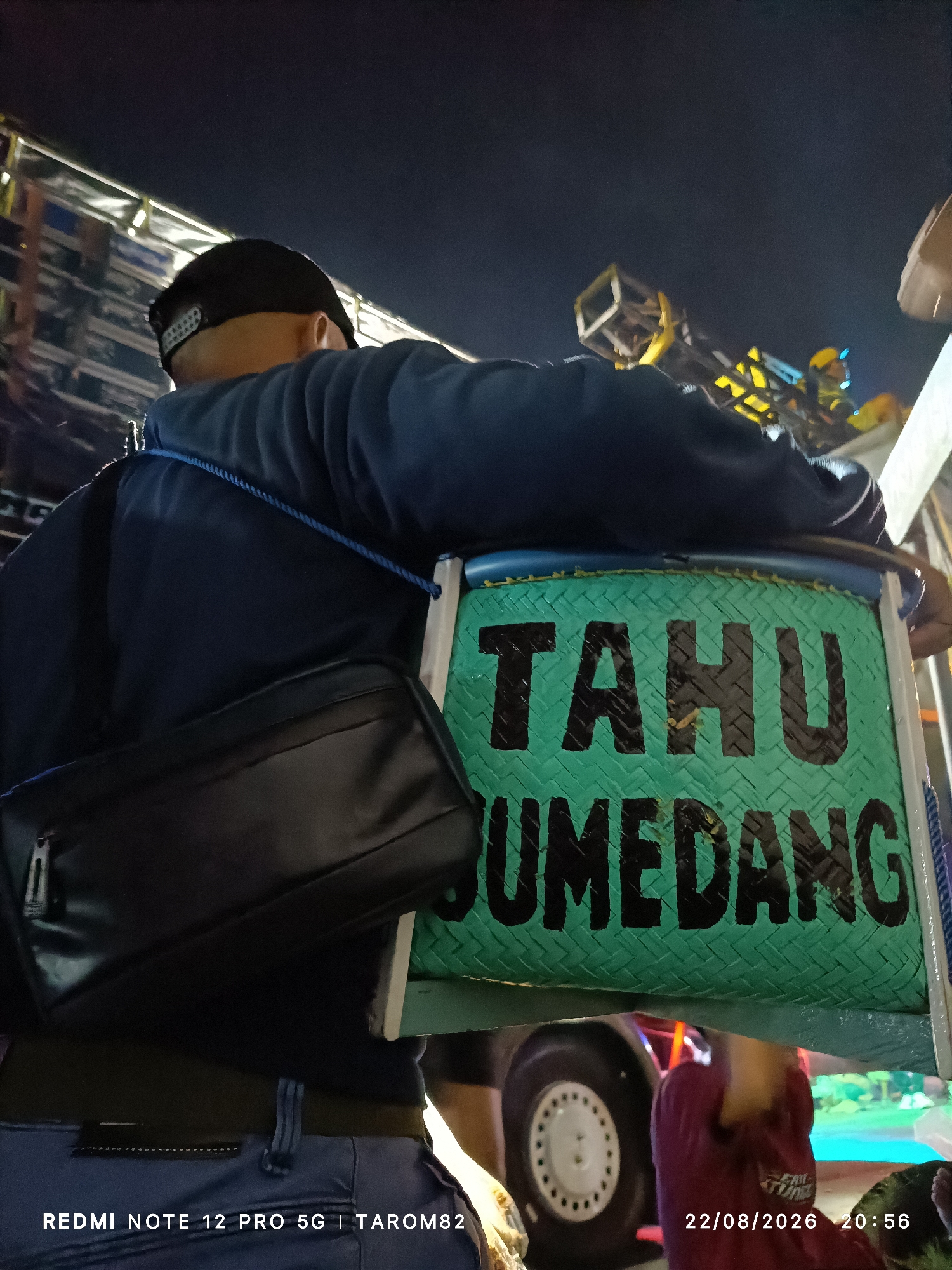 In Mundusewu, Indonesia, a person wearing a blue jacket and a black baseball cap is seen from behind, carrying a green woven basket with "TAHU SUMEDANG" written in black on it. A black sling bag is slung across their body. The background is dark, suggesting it's nighttime, with some blurred lights and structures visible. The image also shows the bottom part of a vehicle with a white wheel. Text at the bottom of the image reads "REDMI NOTE 12 PRO 5G | TAROM82 22/08/2026 20:56".