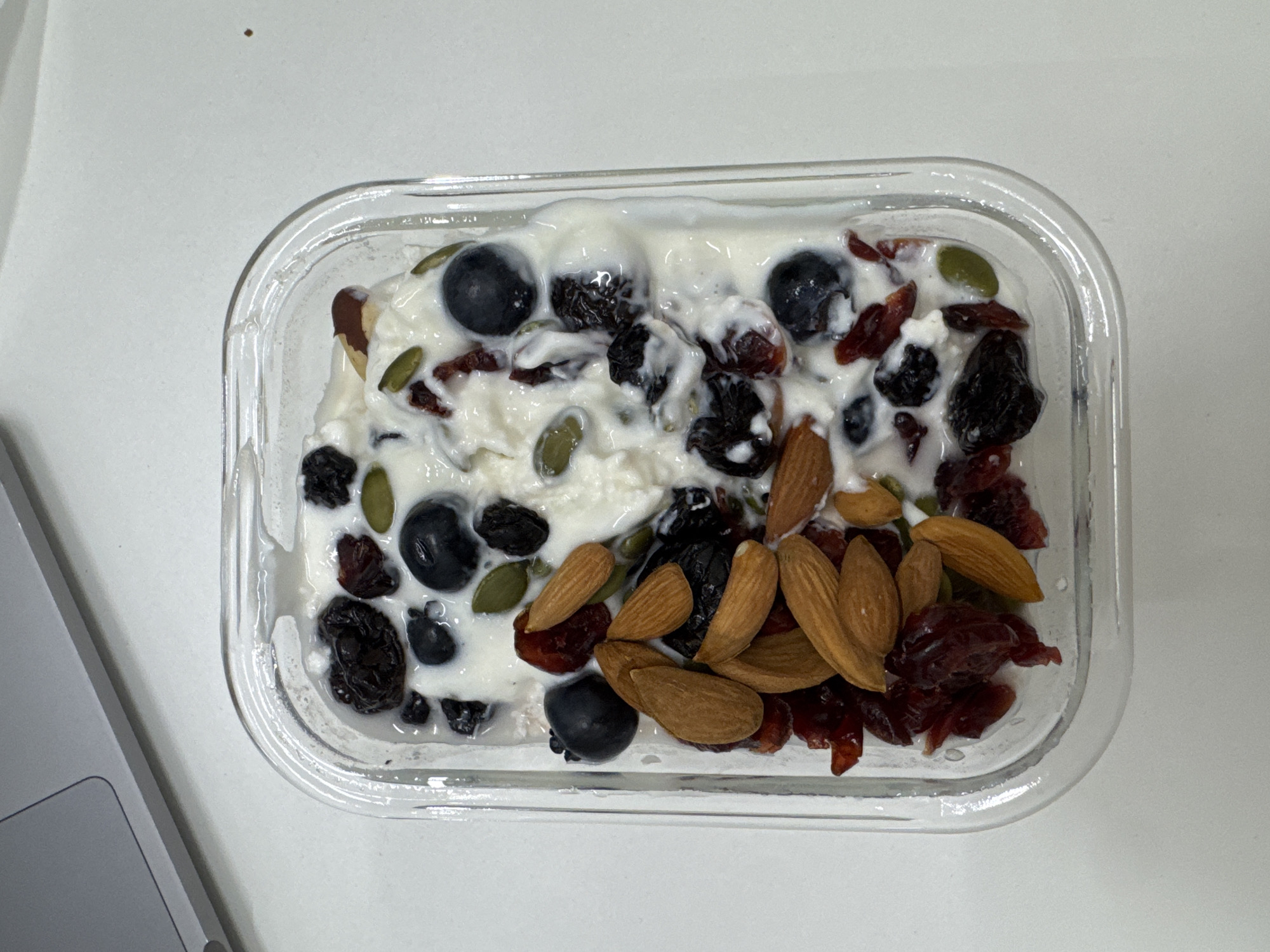 A close-up, overhead shot displays a healthy snack or meal contained within a clear, rectangular glass dish with rounded corners. The dish holds a creamy white base, resembling yogurt or cottage cheese, densely topped with a variety of ingredients. These toppings include dark blue blueberries, reddish dried cranberries, dark, shriveled dried berries (possibly raisins or dried cherries), vibrant green pumpkin seeds, and light brown whole almonds. The mixture of toppings is spread across the surface of the creamy base.
The glass container is resting on a plain white, possibly matte, surface, suggesting an indoor setting like a desk or table. In the bottom-left portion of the frame, a silver-colored laptop is partially visible, indicating a personal or work environment. The lighting is bright and uniform, but there are no cues to determine the time of day or specific weather conditions. No individuals are present in the image, and no specific activity or event beyond the presentation of the prepared food is depicted. There is no visible text anywhere in the image. While the location context is provided as Dubai, United Arab Emirates, there are no visual elements within the image itself to confirm this geographical location.