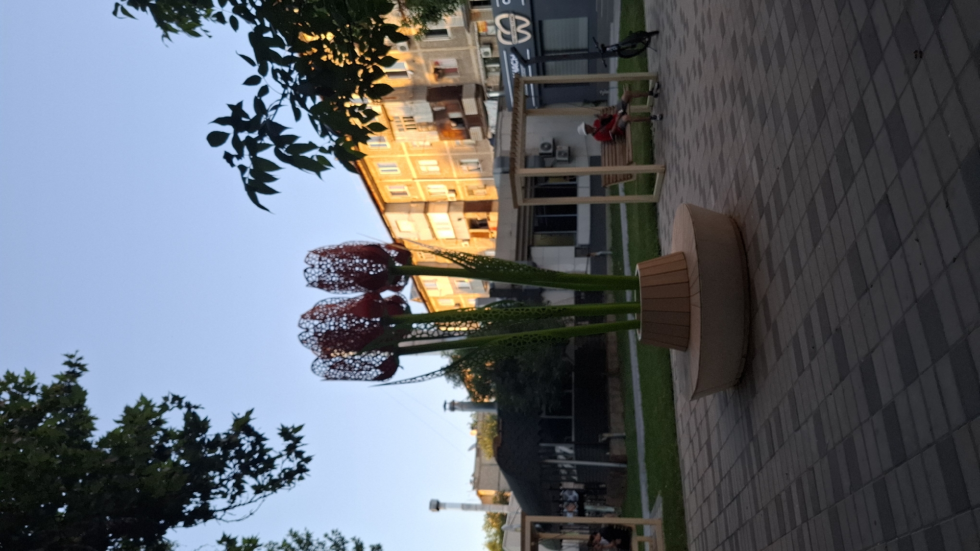 The image depicts an outdoor public space, likely a park or a pedestrian area, bathed in the warm glow of late afternoon or early evening sun. In the foreground, a paved walkway composed of rectangular gray and beige tiles leads the eye towards a prominent art installation of three large, stylized tulips. These tulips are crafted from metal with intricate, perforated red petals and green, segmented stems. They stand in a circular, wooden planter.
To the right of the art installation, a man wearing a red shirt and a white hat is seated on a wooden swing. A bicycle is parked beside the swing. In the background, a multi-story apartment building with a light-colored facade is visible, its windows catching the sunlight. Adjacent to the building is a row of shops, one of which has a sign with a dental logo. Trees with dark green foliage frame the top and sides of the image, casting shadows on the scene. A hint of a dark, covered structure, possibly a cafe or shelter, can be seen further in the background. The sky is a clear, pale blue, indicating fair weather. This scene is located in the city of Shymkent, Kazakhstan.