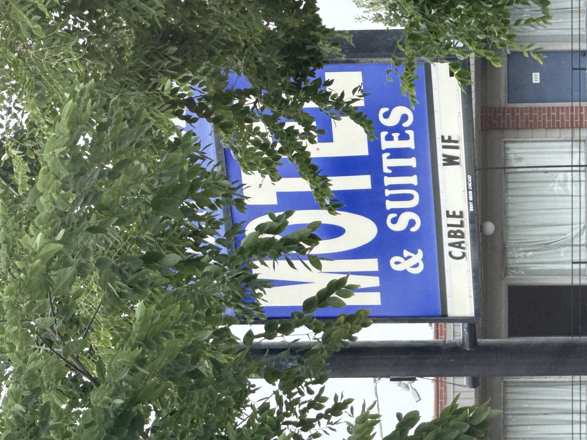 A blue and white sign for a motel reads "MOTEL & SUITES" in large white letters. Below this, smaller white letters spell out "CABLE WIF". Underneath this, in even smaller print, it says "BEST NEON CHICAGO". The sign is partially obscured by green foliage from a tree in the foreground. In the background, a building with windows and a blue door with the number "220" on it is visible. The image appears to be taken outdoors, possibly in a city setting, given the presence of the motel sign and building.