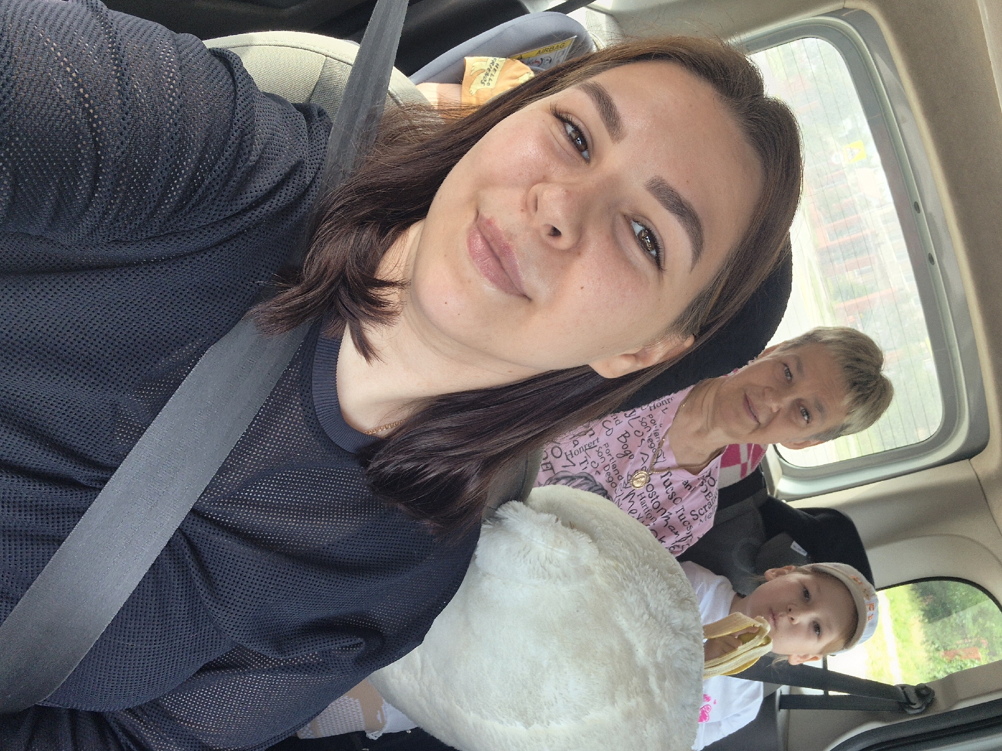 In this selfie taken from the back seat of a car, a woman with brown hair, wearing a black mesh shirt and a seatbelt, is smiling at the camera. Behind her, a woman with short blonde hair and a pink t-shirt is visible, and to the right, a young boy in a white t-shirt and a baseball cap is eating a banana. The interior of the car appears to be a typical family vehicle. The background seen through the car window shows trees and buildings, suggesting an urban or suburban environment. The image does not provide specific visual cues that can definitively place the location within the city of Chaikovsky, Russia.