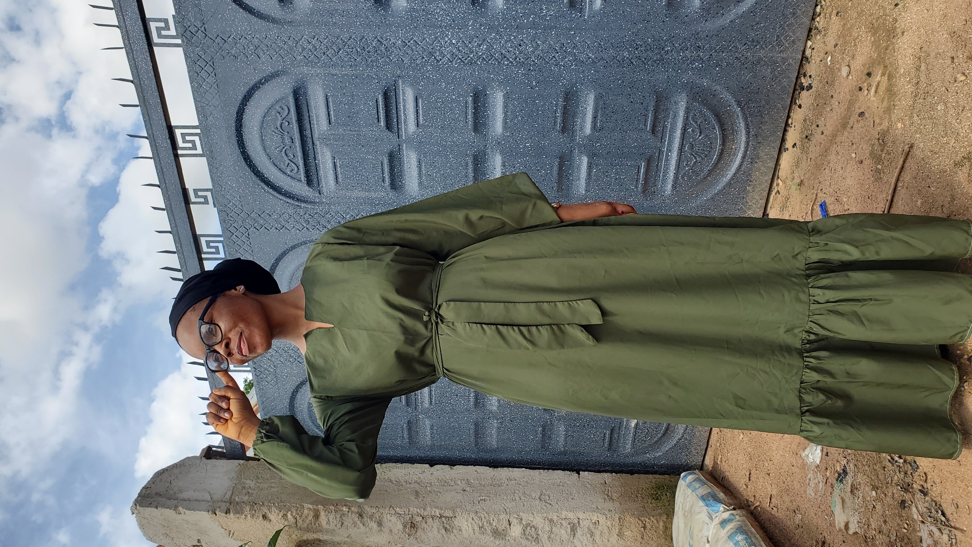 A young Black woman wearing glasses and a black hijab stands outdoors in front of a grey metal gate. She is wearing a long, olive-green dress with long, wide sleeves and a ruffled hem. The dress is cinched at the waist with a matching fabric belt. Her right hand is raised, and she appears to be adjusting her glasses. The background consists of a stone structure on the left, the grey metal gate with decorative patterns, and a cloudy sky above. The ground is sandy and appears to be a dirt road. The scene is likely in Jalingo, Nigeria, a city known for its residential areas and gates.