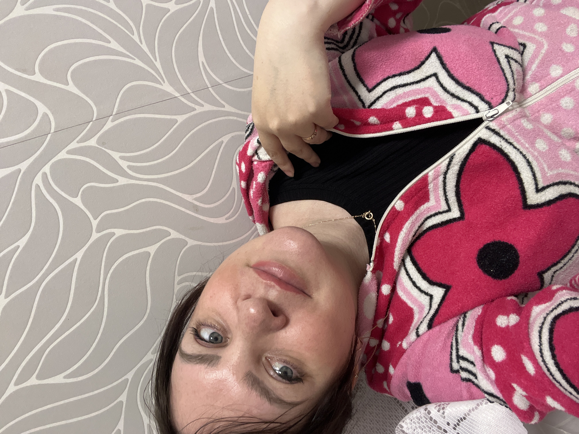 A woman with fair skin, dark eyebrows, and dark hair, wearing a black top and a pink floral-patterned zip-up jacket, looks directly at the camera. A gold necklace and ring are visible. The background is a grey wall with a white, repeating leaf-like pattern. A portion of what appears to be a white lace tablecloth is visible in the bottom right corner. The location is Perm, Russia.
