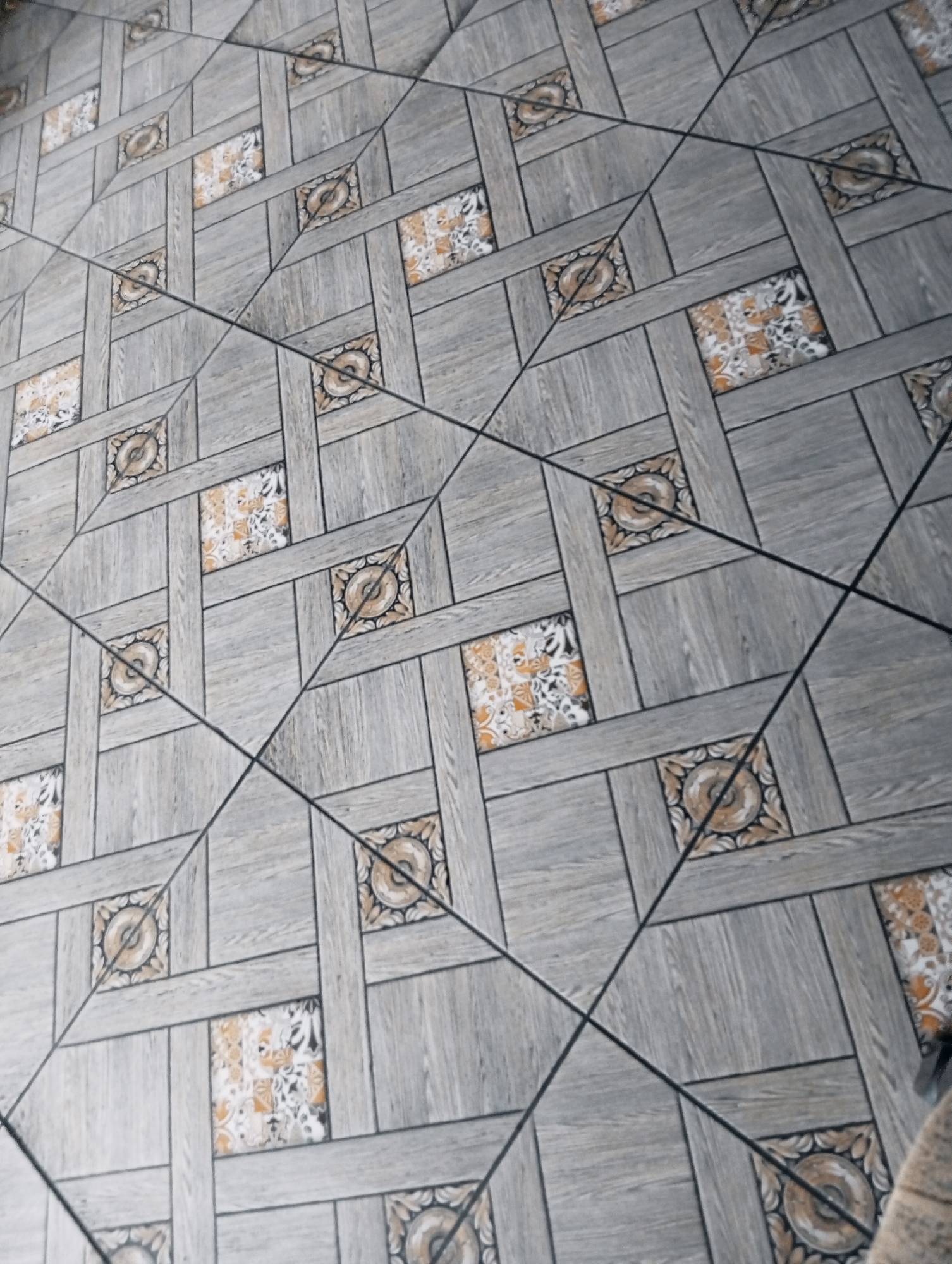 The image shows a floor with a patterned tile design. The tiles are predominantly gray with a wood-like texture, accented by smaller square insets featuring floral or geometric patterns in shades of yellow and white. The pattern repeats across the floor, creating a grid-like appearance. Dark lines run diagonally across the surface, likely grout lines between the tiles. A portion of what appears to be a light brown rug is visible in the lower right corner. There is no information to infer the location of this image in the city of Poltava, Ukraine.