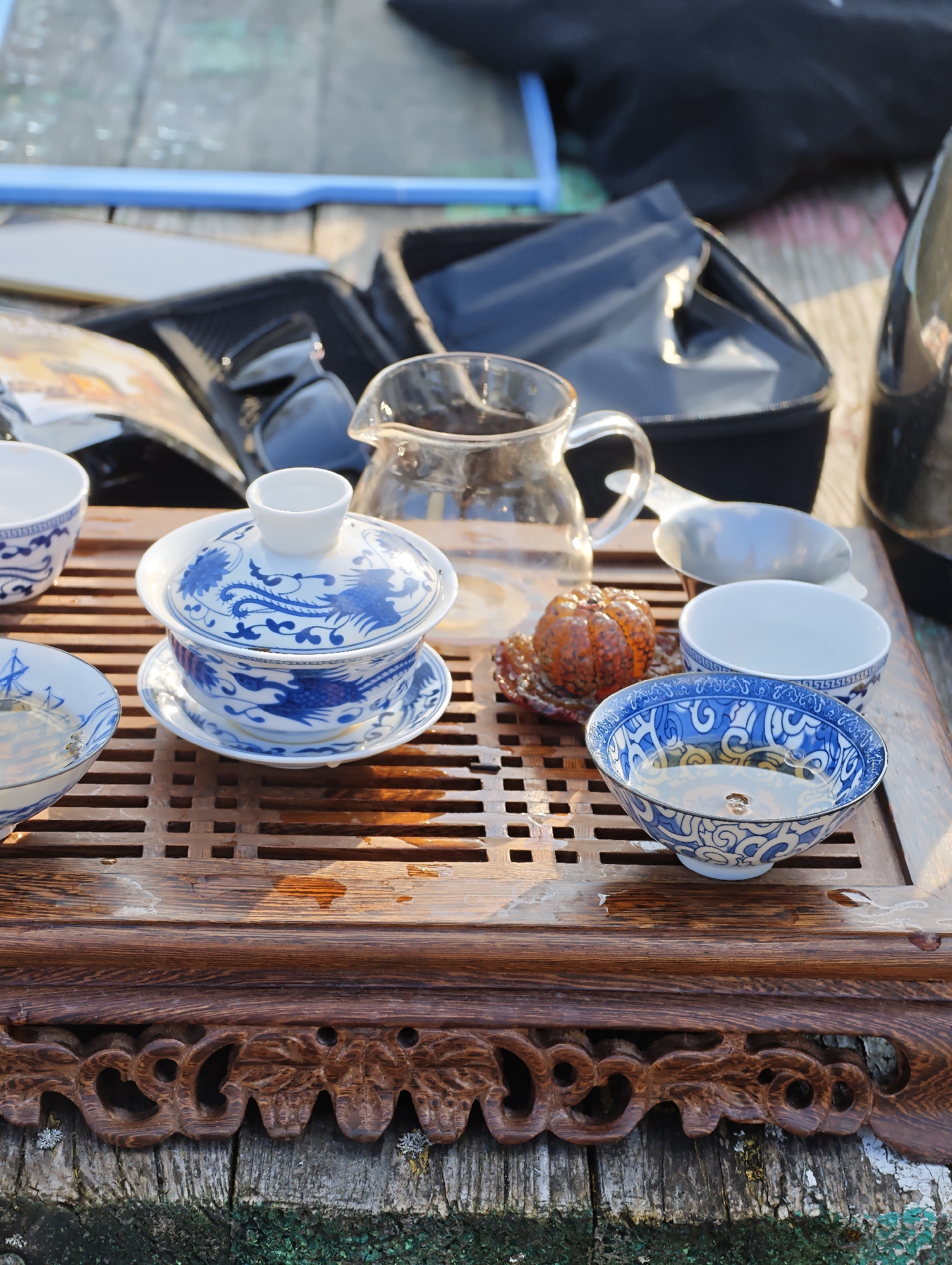 The scene, situated outdoors in Парэчча, Belarus, depicts a collection of tea ceremony items arranged on a dark, carved wooden tea tray. The tray features a slatted top, upon which multiple water droplets are visible, and rests on weathered, grey wooden planks showing patches of green moss or discoloration and white paint residue.
Centrally located on the tray is a white porcelain gaiwan, comprising a lidded cup and saucer, all intricately decorated with blue traditional patterns, including a stylized dragon or phoenix motif. To the left, a white porcelain bowl with a blue ship design holds a clear, pale liquid. To the right, a clear glass fairness pitcher (gongdao bei) stands, partially filled with liquid. Adjacent to the pitcher are two additional small white porcelain cups, one featuring a blue geometric pattern and containing a clear liquid, and the other plain white. A small, orange, textured spherical object, possibly dried citrus peel or a tea cake, rests on a reddish-brown, textured coaster behind the foreground patterned bowl. A small, silver-colored scoop or strainer is positioned behind the fairness pitcher. In the blurred background, dark fabric items, possibly bags or cases, and a partial blue object are discernible. No individuals are present within the frame.