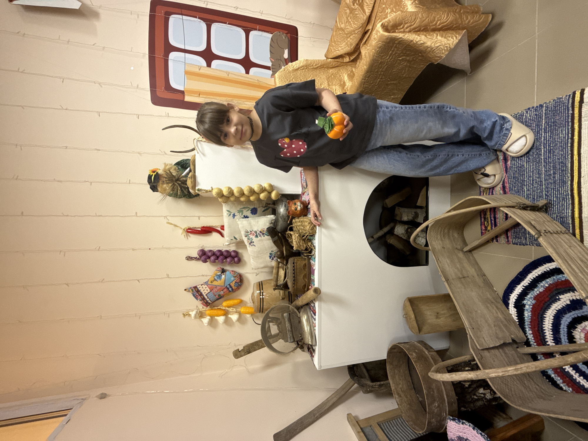 A child stands next to a white stove in a room designed to resemble a rural Russian interior, possibly in a museum or historical exhibit in Rusanovo, Russia. The stove, which features an opening showing stacked firewood, is positioned against a wall decorated with various traditional household items, including strings of garlic, dried peppers, and other implements, giving the impression of a rustic kitchen. The child, wearing a dark t-shirt with a heart-shaped design and jeans, holds small, orange-colored objects, possibly decorative pumpkins. Behind the child, a window painted on the wall shows a landscape scene. A woven rug lies on the floor next to a wooden chair. A golden-colored fabric is draped in the background.