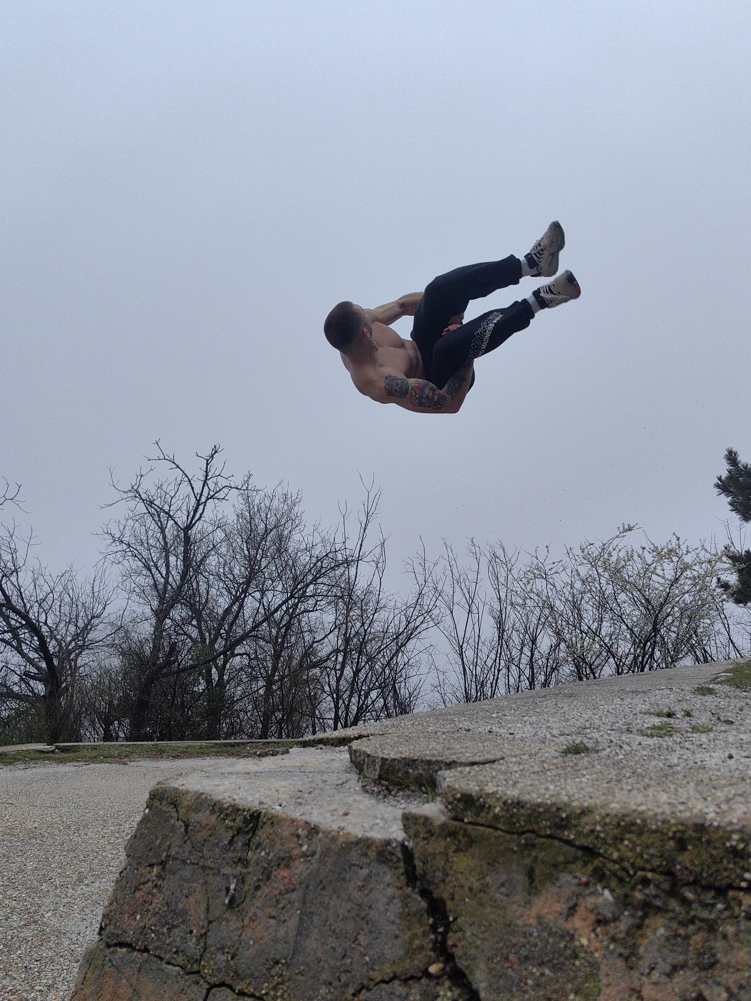 A shirtless man wearing black pants with white writing on the lower leg, socks, and sneakers is performing a backflip in mid-air. He is above a cracked concrete wall with vegetation behind it, including bare trees and trees with budding leaves. The sky is overcast. The location is in Varna, Bulgaria, based on the presence of the Asparuhov Bridge in the background (not fully visible in the provided image). The man appears to be doing calisthenics or parkour.