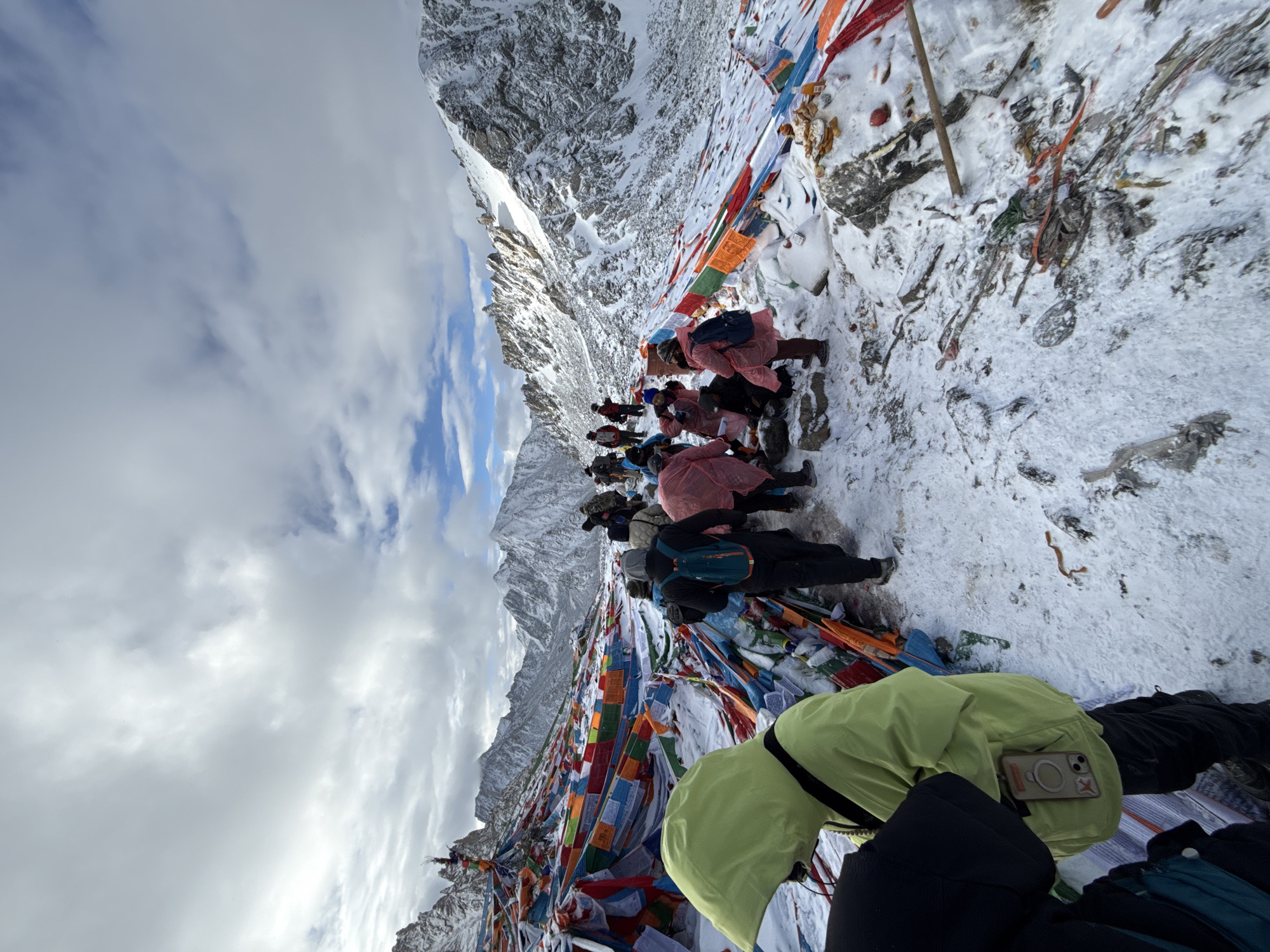 This high-angle, wide-shot photograph captures a group of pilgrims engaged in a spiritual ritual on a snowy mountain pass, likely near Parga town, China, characterized by an abundance of colorful Tibetan prayer flags. The scene is dominated by a majestic, snow-capped mountain range under a partly cloudy sky, with patches of blue visible.
In the foreground and middle ground, several individuals, dressed in warm winter clothing including jackets and rain ponchos (in shades of pink, lime green, and dark colors), are seen prostrating or bowing low on the snow-covered ground. Many wear backpacks, suggesting a long journey or pilgrimage. The people are arranged in a line, extending into the distance along the mountain ridge, implying a procession or collective prayer. The vibrant prayer flags, or lungta, are strung across the landscape, draping over rocks and extending along the path, adding a strong cultural and spiritual dimension to the rugged environment.
The ground is rocky and uneven, covered with fresh snow, and interspersed with debris and offerings, such as small fabric pieces, sticks, and other items. The weather appears cold and overcast, though light conditions suggest daytime, possibly late morning or early afternoon. One person in the immediate foreground, wearing a lime-green jacket, has an iPhone with a distinct MagSafe wallet or grip (featuring an "X" or cross symbol) visible in a back pocket. The overall atmosphere conveys a sense of deep devotion and perseverance amidst the challenging, high-altitude environment.