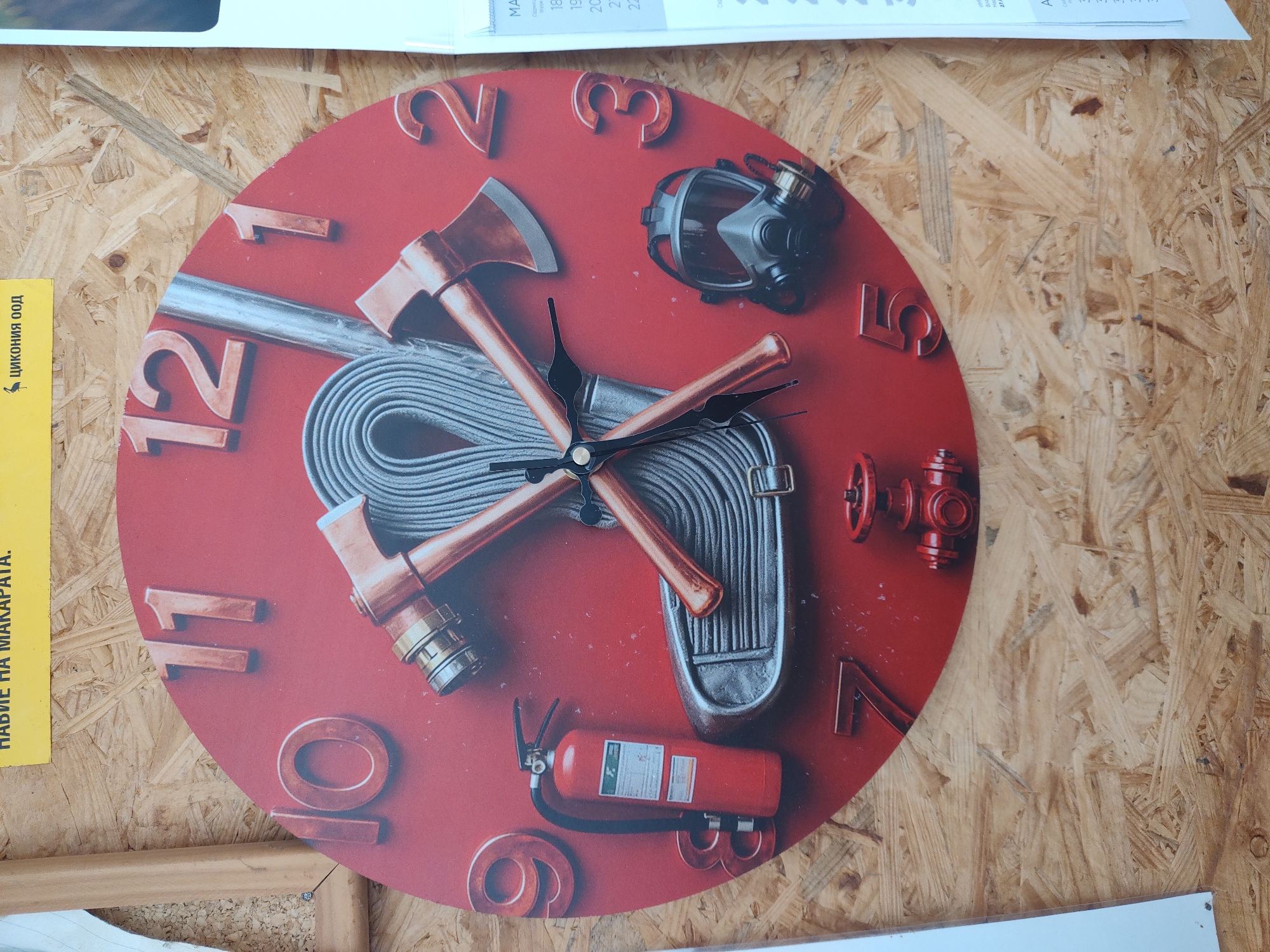 The image displays a close-up, eye-level, full shot of a circular wall clock with a red face, designed with a fire-fighting theme. The clock face features large, raised, metallic-looking numbers for each hour, from 1 to 12. The hands of the clock are thin and black, pointing to approximately 2:45. The clock's design incorporates various firefighting equipment: two axes crossed in the center, a coiled hose, a fire extinguisher on the left side, a fire hydrant valve at the bottom, and a gas mask on the right side. The clock is mounted on a textured wooden background, likely a wall made of OSB (Oriented Strand Board). The lighting suggests an indoor environment, possibly a workshop or a themed exhibition space. There are no people visible in the image, nor any discernible elements that would specifically place it within the city of Tervel, Bulgaria. The scene is static and focused solely on the clock and its immediate surroundings.