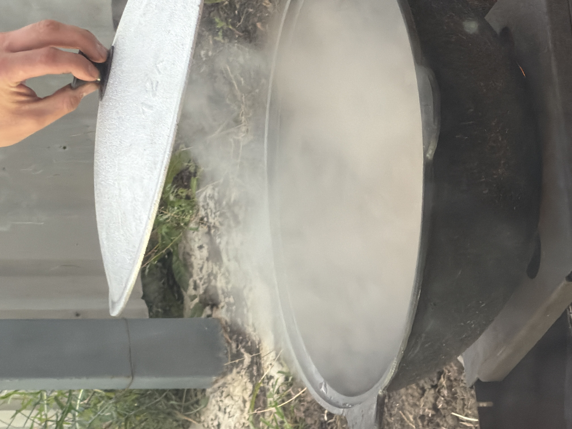 The image shows a close-up, rotated view of a large, dark pot with a light-colored lid being held slightly ajar by a person's hand. Steam is billowing out from the opening of the pot, suggesting that something is being cooked or heated inside. The pot appears to be made of cast iron and has a textured, dark exterior. The lid is textured and appears to be made of a lighter material, possibly aluminum or a similar metal. The hand holding the lid is lightly gripping a knob or handle on its edge.
The scene is set outdoors, with glimpses of dirt, dry grass, and possibly some vegetation around the pot. A gray cylindrical object, possibly part of a structure or support, is visible in the lower left portion of the frame. The lighting suggests it is daytime, but the weather is not explicitly discernible. The overall impression is one of food preparation, likely in an outdoor setting such as a campsite or a backyard. There is no visible text in the image. The setting hints at a rustic or outdoor culinary activity.
