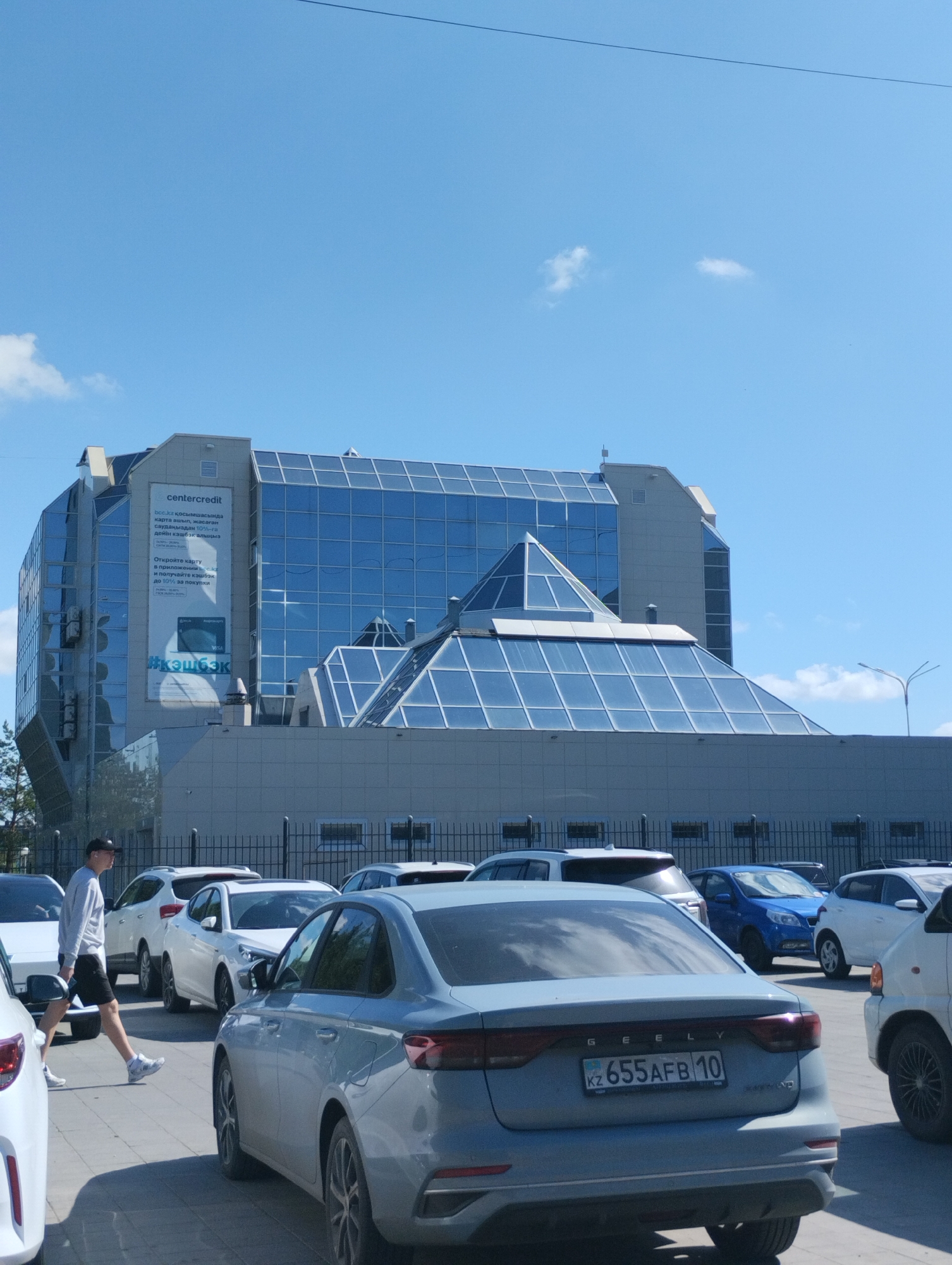 An outdoor medium shot captures a bright, sunny day in Kostanay, Kazakhstan, with a modern, multi-faceted building dominating the background. The building features extensive blue glass panels and a pyramidal glass roof structure. A prominent sign on the building reads "centercredit" and contains text in Cyrillic script, along with advertisements for a credit card. In the foreground, several cars are parked. A person wearing a light-colored long-sleeved shirt, black shorts, a baseball cap, and sneakers is walking across the paved area. The ground is made of interlocking concrete pavers. The sky is clear blue with a few small white clouds.