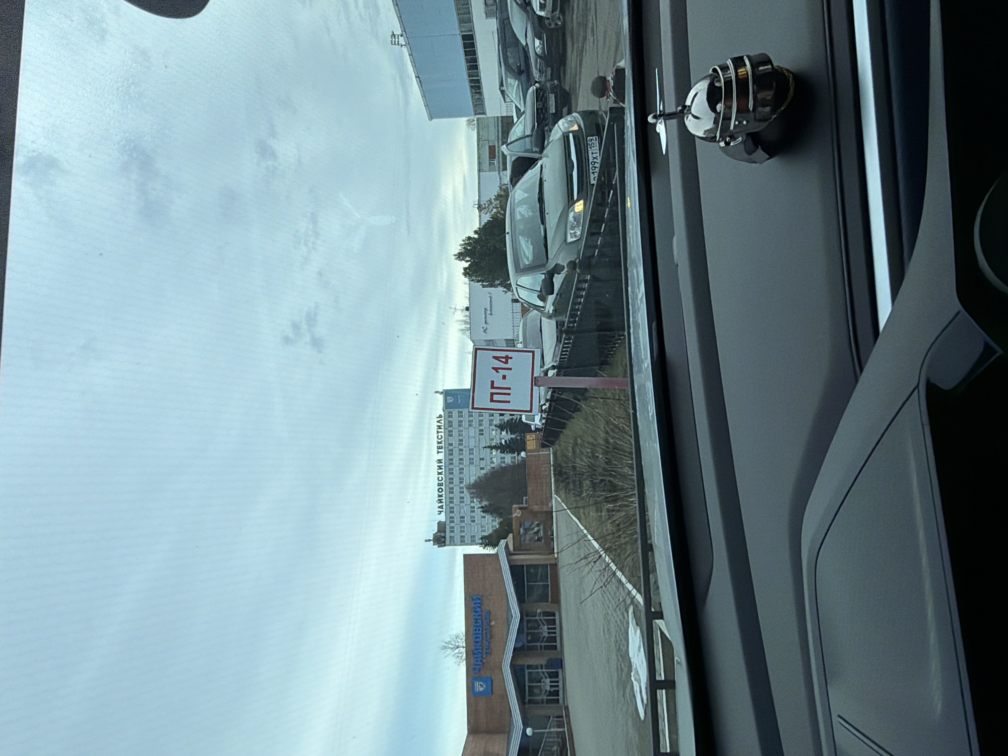 The image, taken from inside a vehicle, shows a street scene in the city of Chaikovsky, Russia. Prominently visible is the building of "Chaikovsky Textile," with the name displayed in blue lettering. A road sign marked "PG-14" is also present. Several cars are parked nearby, including a dark-colored car with license plate "A997X7159." The sky is overcast. Inside the vehicle, a chrome-colored decorative object is affixed to the dashboard.