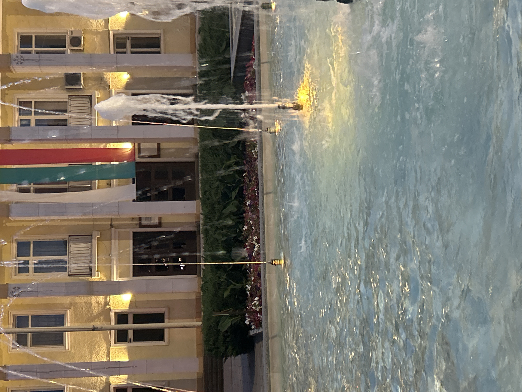 The image shows a fountain in front of a yellow building with columns and arched windows. There are also some flags hanging from the building. The fountain is spraying water into the air, and the water is reflecting the light from the building. The scene appears to be in Parvomay, Bulgaria. The building is likely a government building or a cultural institution, given the flags and the formal architecture. The fountain and the surrounding flowers suggest a public space designed for recreation and beautification.