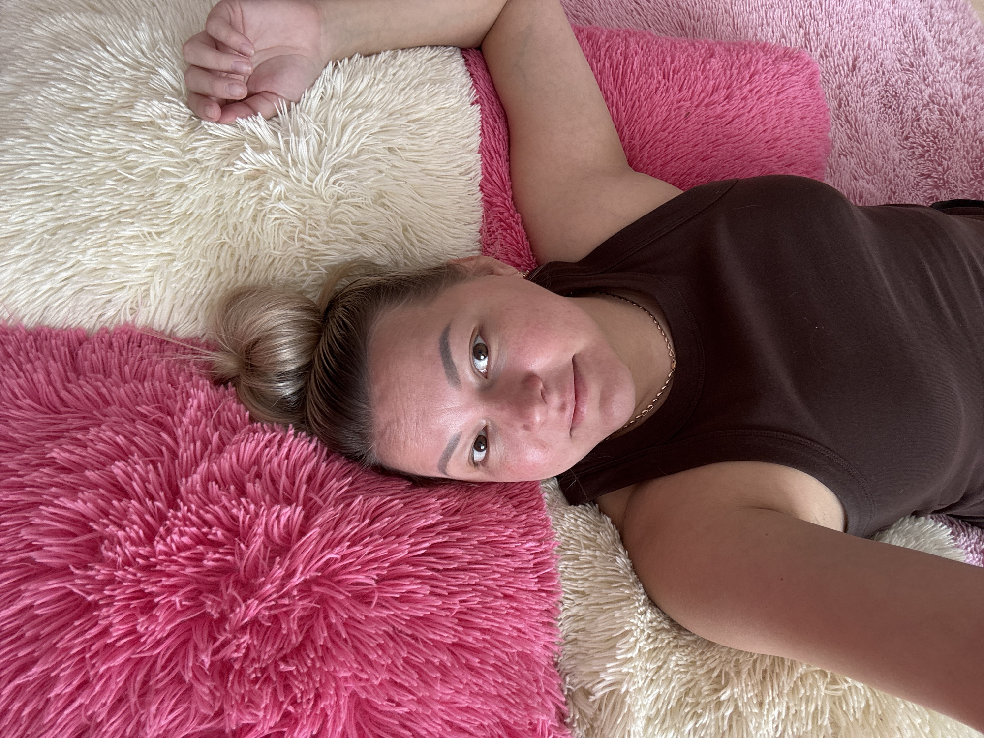 A female subject lies supine on a surface composed of alternating cream-colored and fuchsia-pink shag-pile cushions or blankets. Her head is positioned on a fuchsia cushion to the lower left, with a cream cushion beneath her right arm and extending to her upper left. She wears a dark brown sleeveless top and a thin gold-colored chain necklace. Her blonde hair is pulled back, and her face, showing some redness on the cheeks, is directed towards the camera with a neutral expression. Her eyes are open, looking forward. Her left arm is bent, with her hand resting above her head on a cream cushion. Her right arm extends towards the lower right of the frame. The setting is an indoor location. No specific city or external geographical identifiers are present within the image.