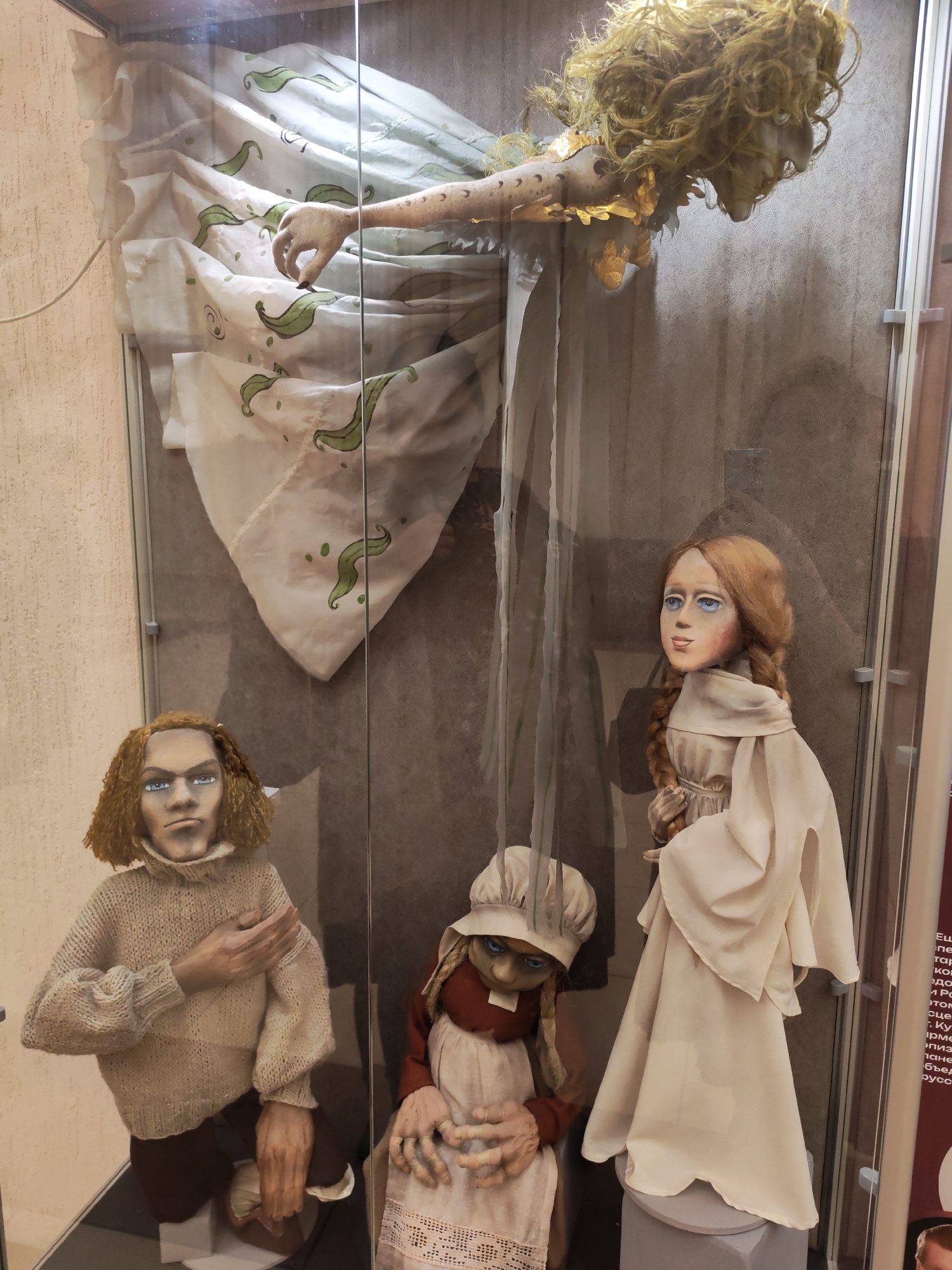 The image displays four intricately crafted dolls or puppets arranged within a glass display case, likely part of a museum or art exhibition. The setting is well-lit, with reflections noticeable on the clear glass, suggesting internal lighting.
At the top of the case, a doll with long, unruly golden hair and a pale face is suspended. Its body is covered in a flowing white garment adorned with a repetitive green, leafy or bean-like pattern. One arm, delicately sculpted and featuring subtle tattoo-like markings, extends downwards, its fingers slightly curled.
Below, on the bottom left, a male doll with short, curly brown hair and a stern expression is positioned in a kneeling stance. He wears a chunky, light beige knitted sweater and dark pants, with one hand placed on his chest. To his right, in the center, a smaller doll, possibly female, is seated. This doll has braided hair, wears a bonnet, a red top, and a white apron, and has striking blue eyes and detailed, wrinkled hands. On the bottom right, a standing female doll with long, braided reddish-brown hair, a pale face, and blue eyes is dressed in a simple, flowing white robe.
The dolls exhibit a distinct, somewhat whimsical yet eerie aesthetic, crafted from materials such as fabric for clothing, painted components for faces and hands, and yarn for hair. The background within the display case is a plain dark grey, providing a stark contrast to the lighter-colored figures. The wall behind the case is textured and light beige. To the far right, a partially visible plaque with Cyrillic text, including words such as "Ещё" (Yet/Still) and parts of "русск" (Russian), indicates explanatory information for the exhibit, consistent with the location context of Izhevsk, Russia.