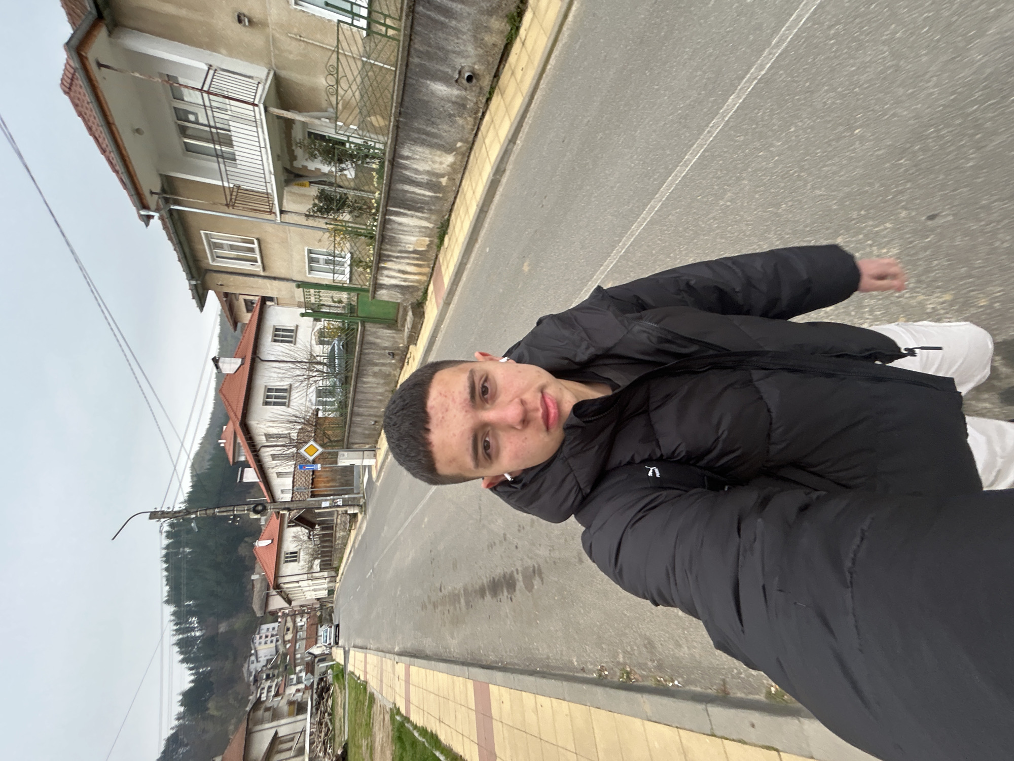 A medium shot captures a young man on a road in Zlatograd, Bulgaria. He is wearing a black puffer jacket and white pants, and facing the camera with a neutral expression. The road runs along a row of buildings, with houses having red-tiled roofs visible in the background, hinting at a residential area. A street lamp and road signs are present, indicating this is a public roadway. The sky is overcast, casting a diffuse light over the scene.