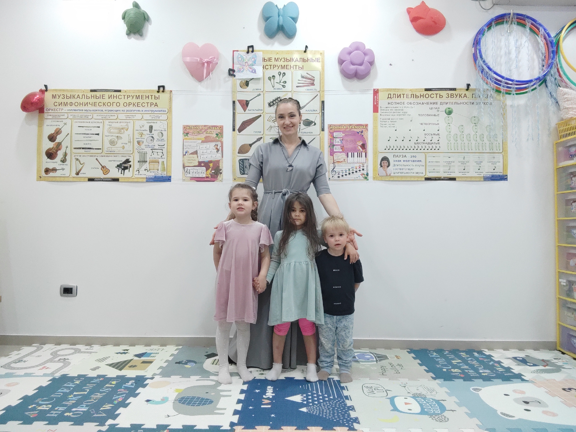 A medium shot captures a woman and three young children standing in front of several educational posters in a classroom setting.
The woman is centrally positioned behind the children, with her hands resting on their shoulders. She has fair skin, light brown hair pulled back, and wears a grey dress with a simple necklace.
The children, two girls and a boy, stand in a row in front of the woman. The girl on the left wears a pink dress and white leggings. The girl in the middle has longer dark hair and wears a light blue dress with pink leggings. The boy on the right has blond hair and wears a black t-shirt with a blue patterned pants. All three children face the camera.
The background wall is adorned with several posters in Russian, likely related to music education. The posters display various musical instruments, note durations, and musical notations. Above the posters are several decorative items, including a green turtle, a pink heart, a blue butterfly, and a purple flower. Hanging on the right side of the frame are several colorful hula hoops.
The floor is covered with interlocking foam tiles featuring alphabet letters, shapes, and illustrations. To the right, a yellow shelving unit can be seen, likely storing classroom materials.
The scene is well-lit, and the overall atmosphere is cheerful and educational. No explicit location information indicates this image was taken in Podgorica, Montenegro.