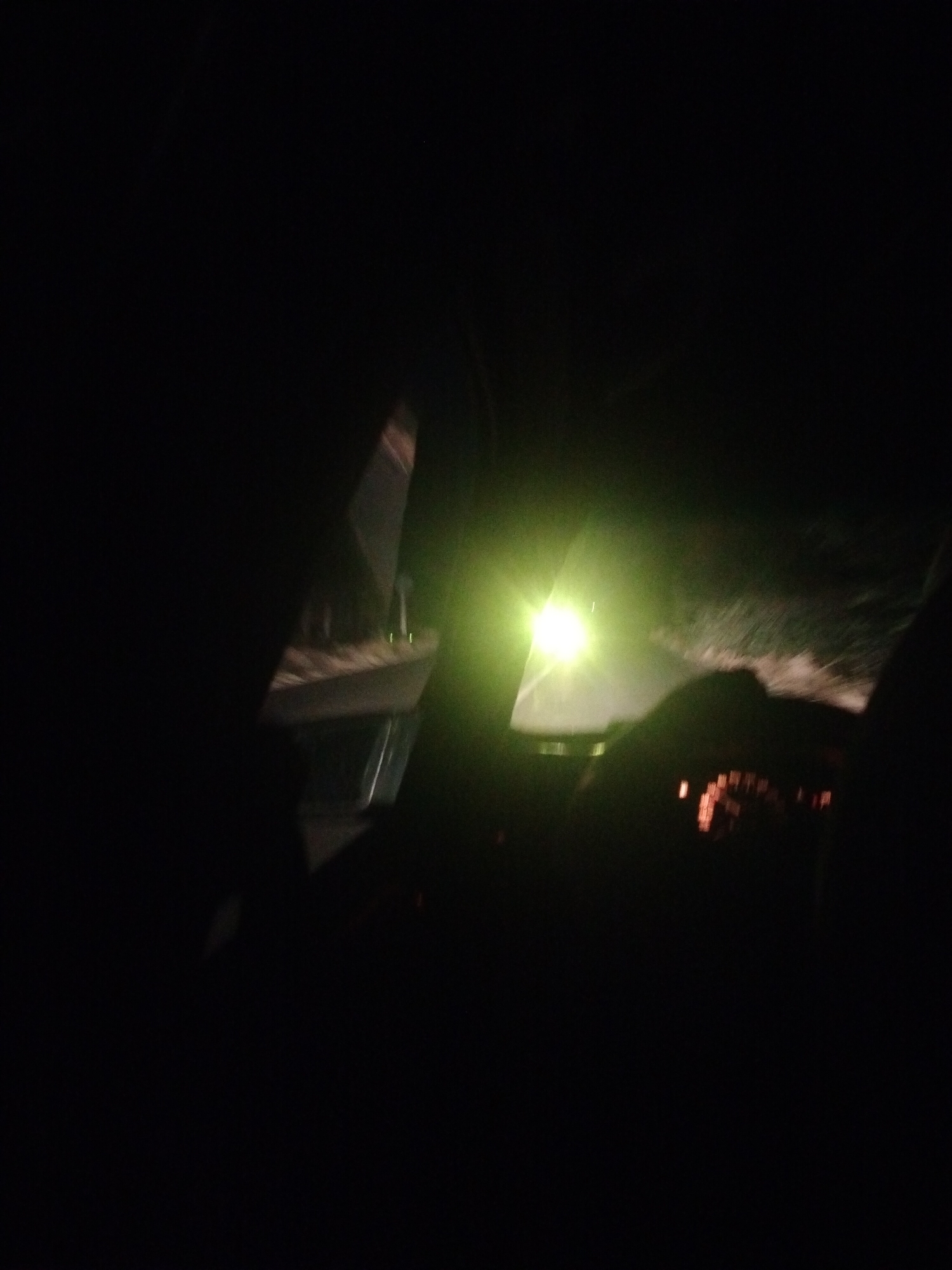 The image depicts a night-time scene from inside a vehicle, likely in or near the city of Qarabulaq, Kazakhstan. The driver's perspective shows a bright, yellow-green headlight beam illuminating a dark, unpaved road or path ahead. The interior of the car is visible, with the dashboard featuring illuminated orange and red indicators. A steering wheel is partially visible in the foreground. The surrounding environment is dark and indistinct, suggesting an rural or undeveloped area outside of city lights. There are no other vehicles or people clearly visible. The overall impression is of a solitary journey through a dark, possibly unlit, landscape.