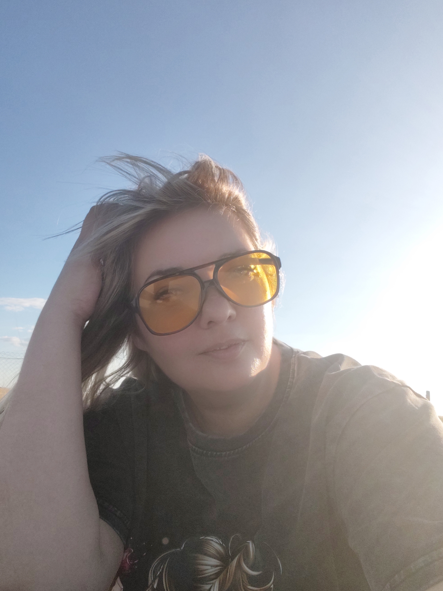 A woman is taking a selfie outdoors with her arm raised and hand in her hair, wearing large orange-tinted aviator sunglasses and a dark t-shirt. The background is a bright, clear blue sky with the sun shining on the right side of the image, creating a lens flare. The woman appears to be outdoors, possibly in a rural or open area, as indicated by the expanse of sky and the lack of urban structures. The location is identified as Nechaiane, Ukraine.