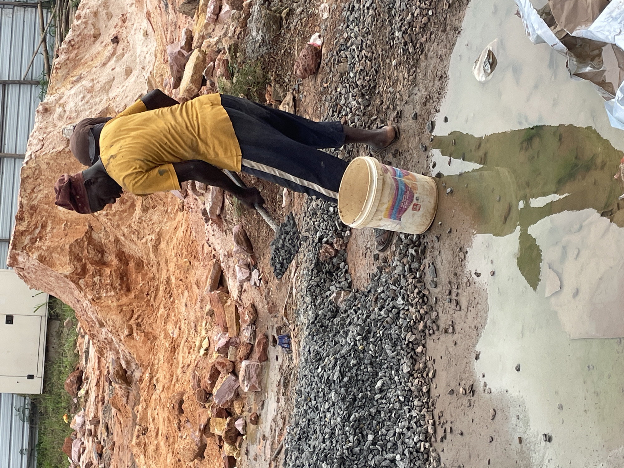 A man, likely a construction worker, is engaged in labor on a construction site in Okinano, Nigeria. He is wearing a yellow t-shirt, dark pants with a white stripe, and a red bandana. He is holding a shovel, appearing to move or gather small dark stones. Beside him, a beige bucket with colorful markings rests on the muddy ground, and a pile of similar stones is visible to his left. In the background, a large mound of reddish-brown earth and rocks dominates the scene. Another individual, only partially visible from the back, is also present on the site. The ground is wet and muddy, with a puddle reflecting the figures and surroundings. Construction materials, including larger rocks and what appears to be a generator or electrical box, are scattered around the site.