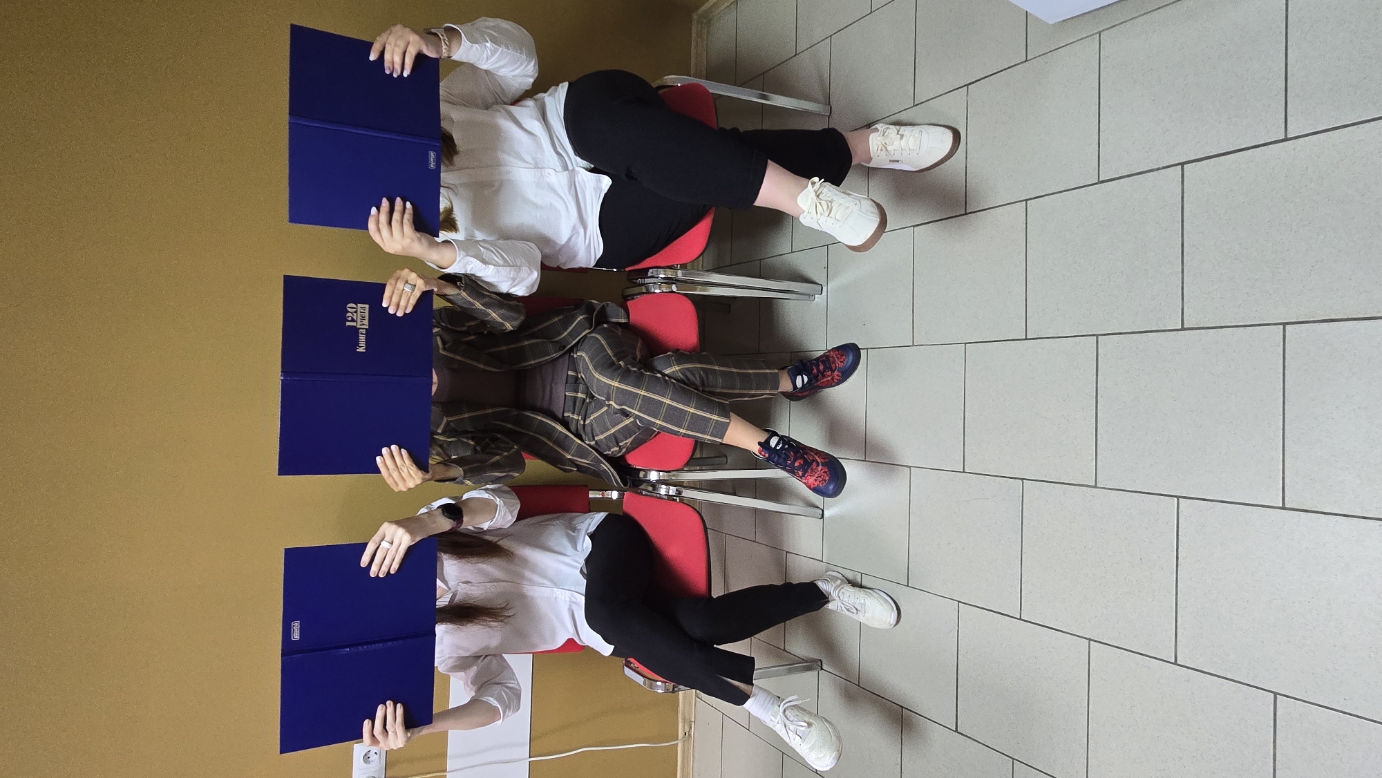 The image shows three people sitting in red chairs, each holding a blue book in front of their face. The people are sitting in a row against a yellow wall. The floor is tiled in light grey. The location appears to be indoors. The specific city is unknown.