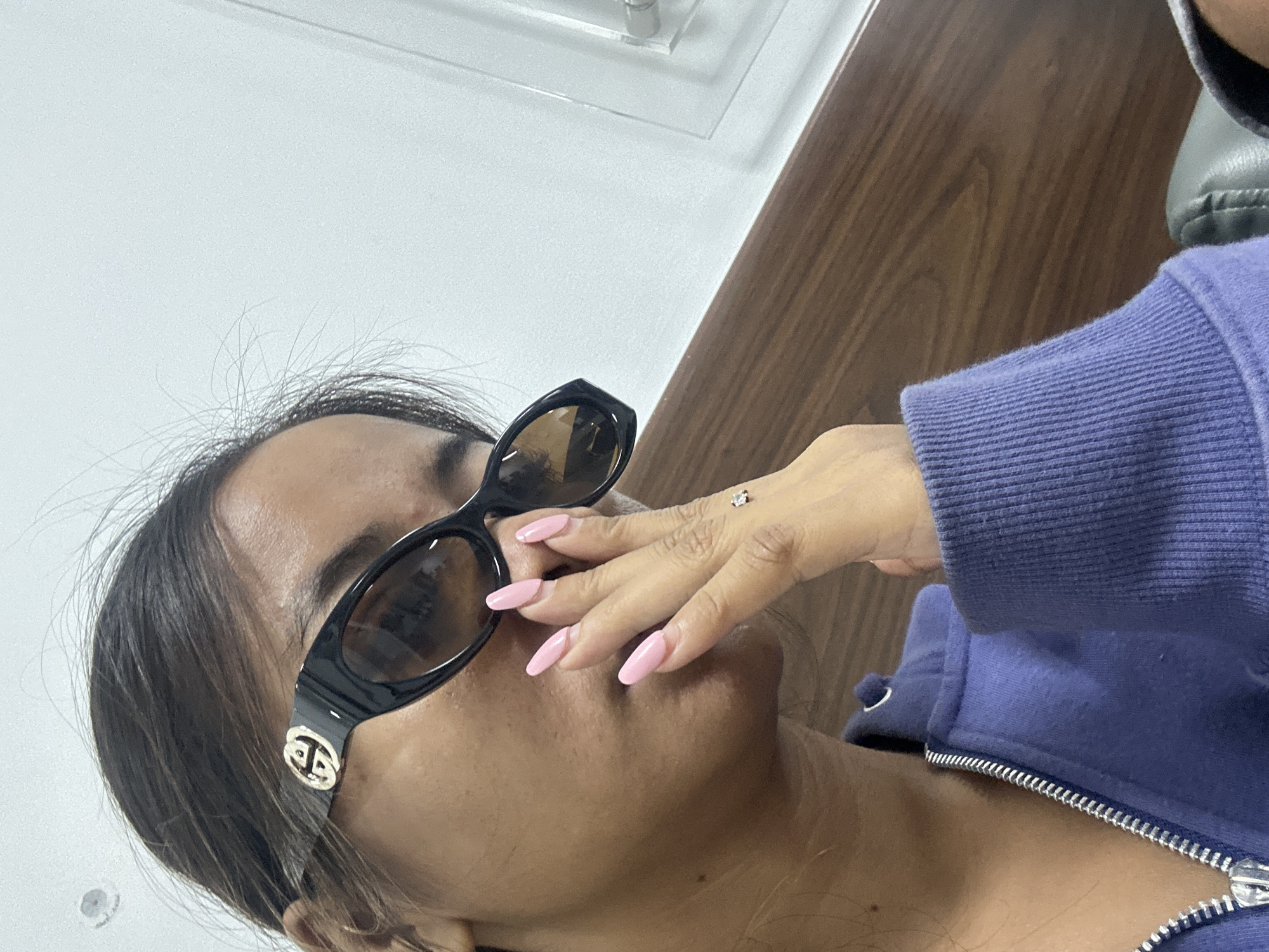 A close-up, medium shot shows a person wearing black sunglasses with gold accents and a purple hooded jacket. The person's face is partially obscured by their hand, which has long, light pink fingernails. A small, square-shaped diamond stud is visible on the back of the person's hand. The background consists of a white wall and a wooden panel. There is no indication of the location being in Kuta, Indonesia.