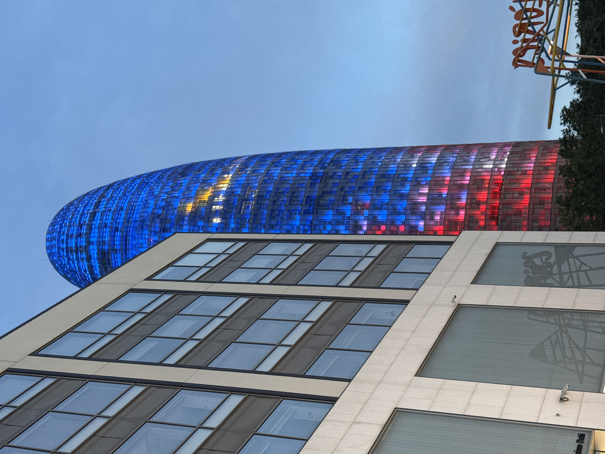 The image captures the distinctive Torre Glòries (formerly Torre Agbar) in Barcelona, Spain, illuminated with vibrant blue and red LED lights against a deep blue twilight sky. The skyscraper's unique bullet-shaped silhouette dominates the right side of the frame, with some yellow/orange lights visible in its upper-middle section. On the left, a modern, multi-story building with a light-colored facade and numerous rectangular, reflective windows occupies the foreground, partially obscuring the base of the Torre Glòries. Reflections of abstract shapes, possibly scaffolding, are visible in the windows of the foreground building. A partial store name, "ssimo Dutti," is reflected in a lower-left window, and surveillance cameras are visible on the building's facade. In the bottom right, a colorful, abstract metal sculpture or sign, possibly part of an art installation, is partially visible, with green tree foliage at the base of the skyscraper. No people are present in the scene. The overall impression is one of modern urban architecture illuminated at dusk.