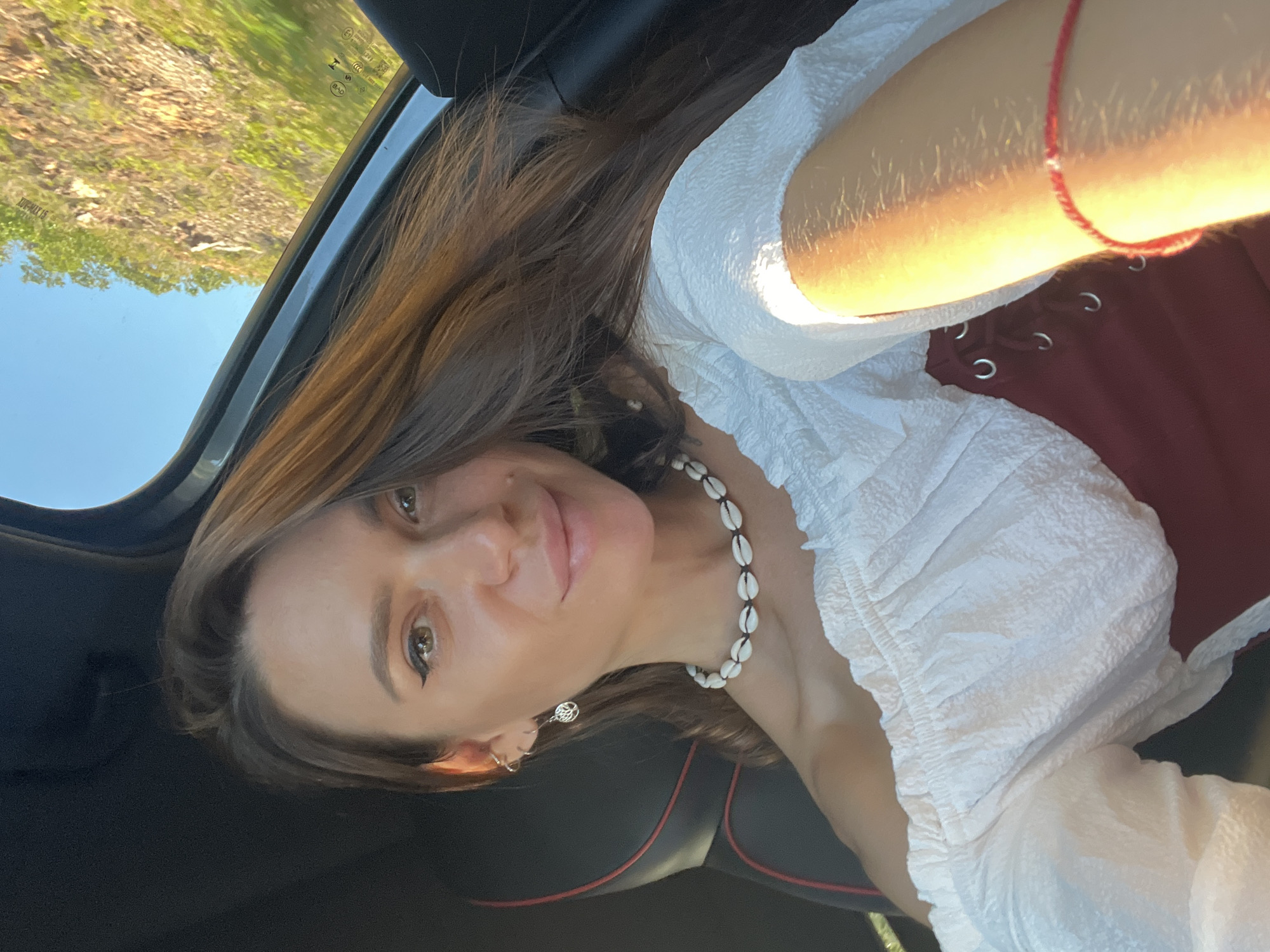 A young woman with long, brown hair is taking a selfie in a car. She is wearing a white, off-the-shoulder top and a dark red corset-style garment underneath. She has a necklace made of small shells and wears small hoop earrings. The car interior is visible, with a black seat and red stitching. Outside the car window, a sunlit hillside with greenery and blue sky can be seen. The lighting suggests it is daytime, possibly late afternoon due to the warm glow.