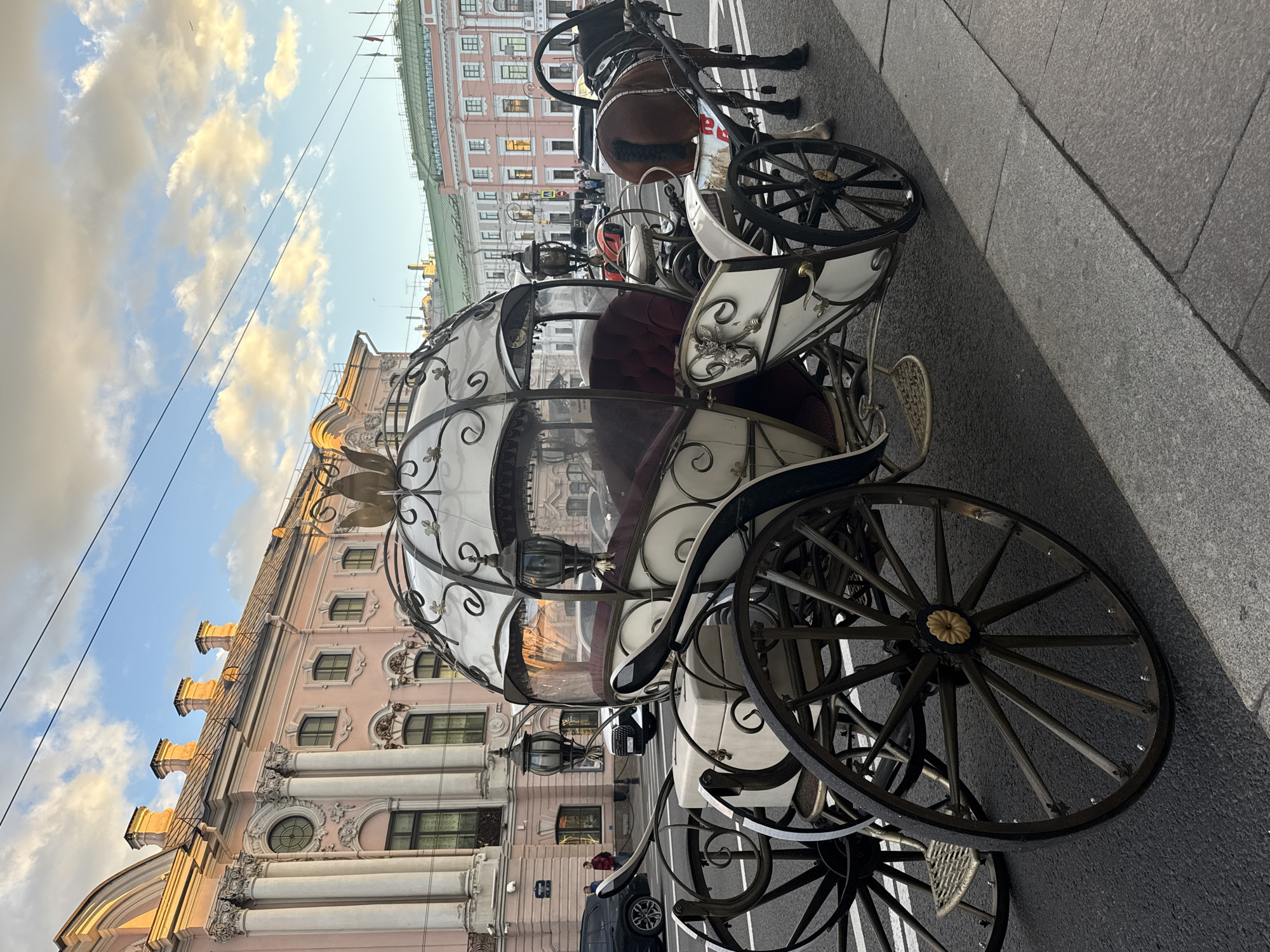 A horse-drawn carriage with a white, ornate body and red velvet seating is parked on a city street. The carriage has large, spoked wheels and is adorned with decorative metalwork. A brown horse, harnessed to the carriage, stands to the right. In the background, classical buildings with pink facades, columns, and arched windows line the street. The sky above is a soft blue with scattered clouds illuminated by the golden light of what appears to be a setting sun. The scene is set on a paved street in Saint Petersburg, Russia, likely near Nevsky Prospect, given the architectural style and the presence of horse-drawn carriages for tourists. Several cars and a few pedestrians are visible in the distance, indicating an urban setting.