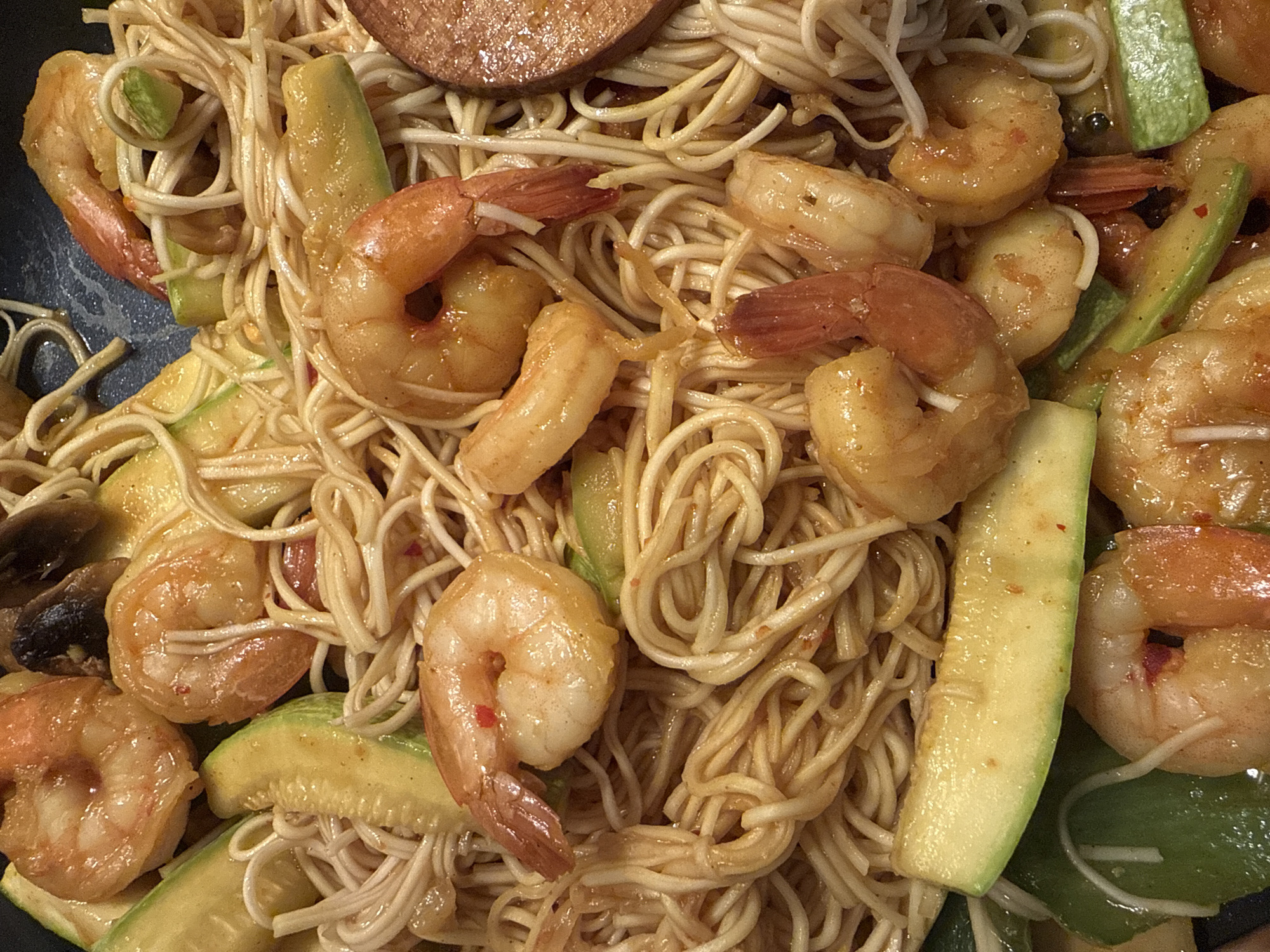 This image shows a close-up of a noodle dish with shrimp and sliced zucchini. The noodles appear to be coated in a light sauce. A single mushroom is visible in the lower left corner, and there is a circular slice of sausage or similar processed meat at the top. The dish is viewed from directly above. The location is not determinable from the visual information.