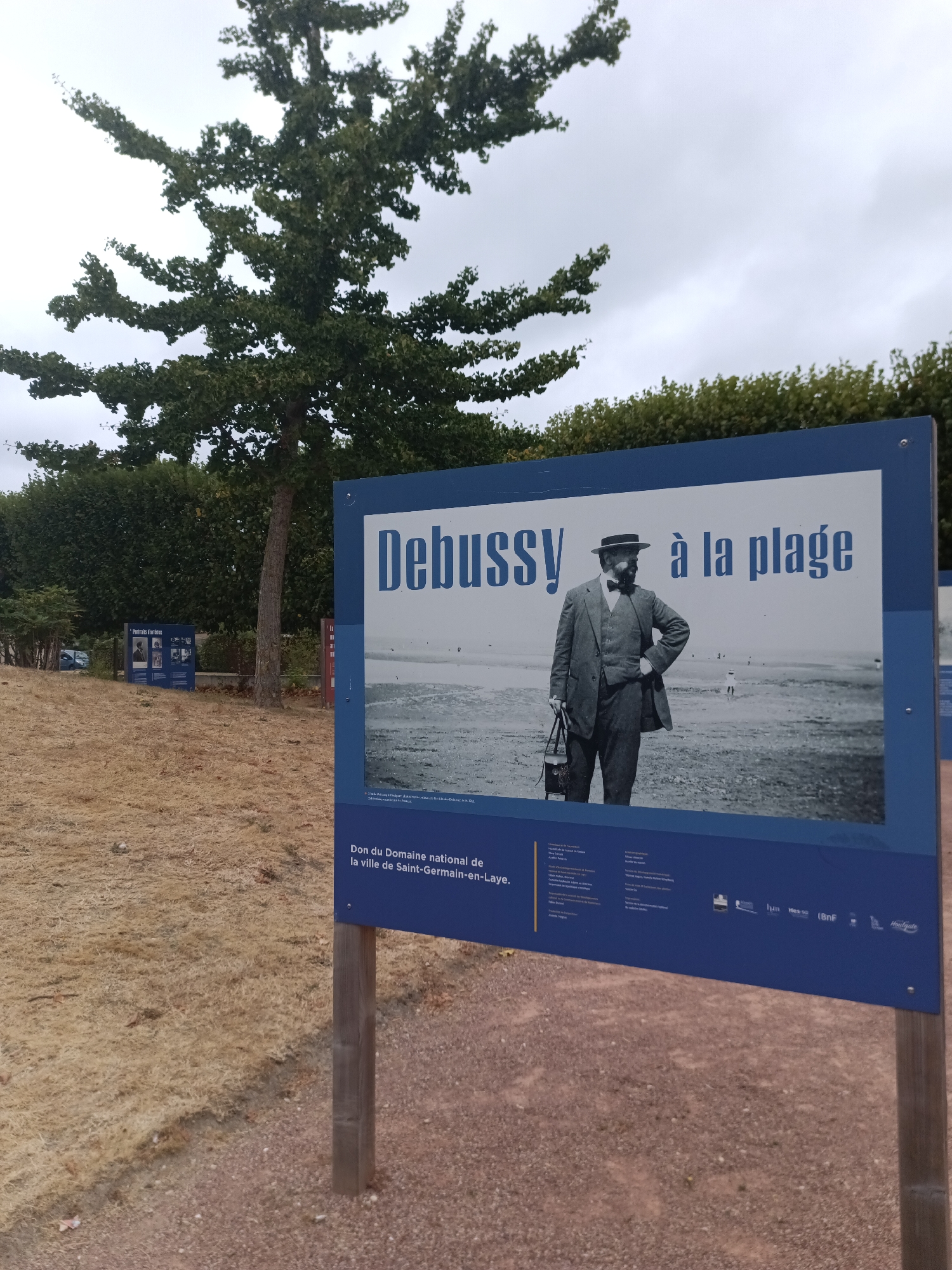 The image shows a large blue informational sign with a black and white photograph of Claude Debussy at a beach. The text on the sign reads "DEBUSSY à la plage." Below the photograph, there is text in French that indicates the sign is a donation from the national domain of the city of Saint-Germain-en-Laye. The sign is positioned outdoors, with a tree and dense foliage in the background. In the distance, there appears to be a body of water and a sandy shore with a few small figures. The ground in the foreground is covered in dry, straw-like grass and some dirt. There are also smaller signs visible behind the main one, and part of another blue sign is visible to the right. The sky is overcast and grey. The overall scene suggests an outdoor exhibition or monument in a park-like setting.