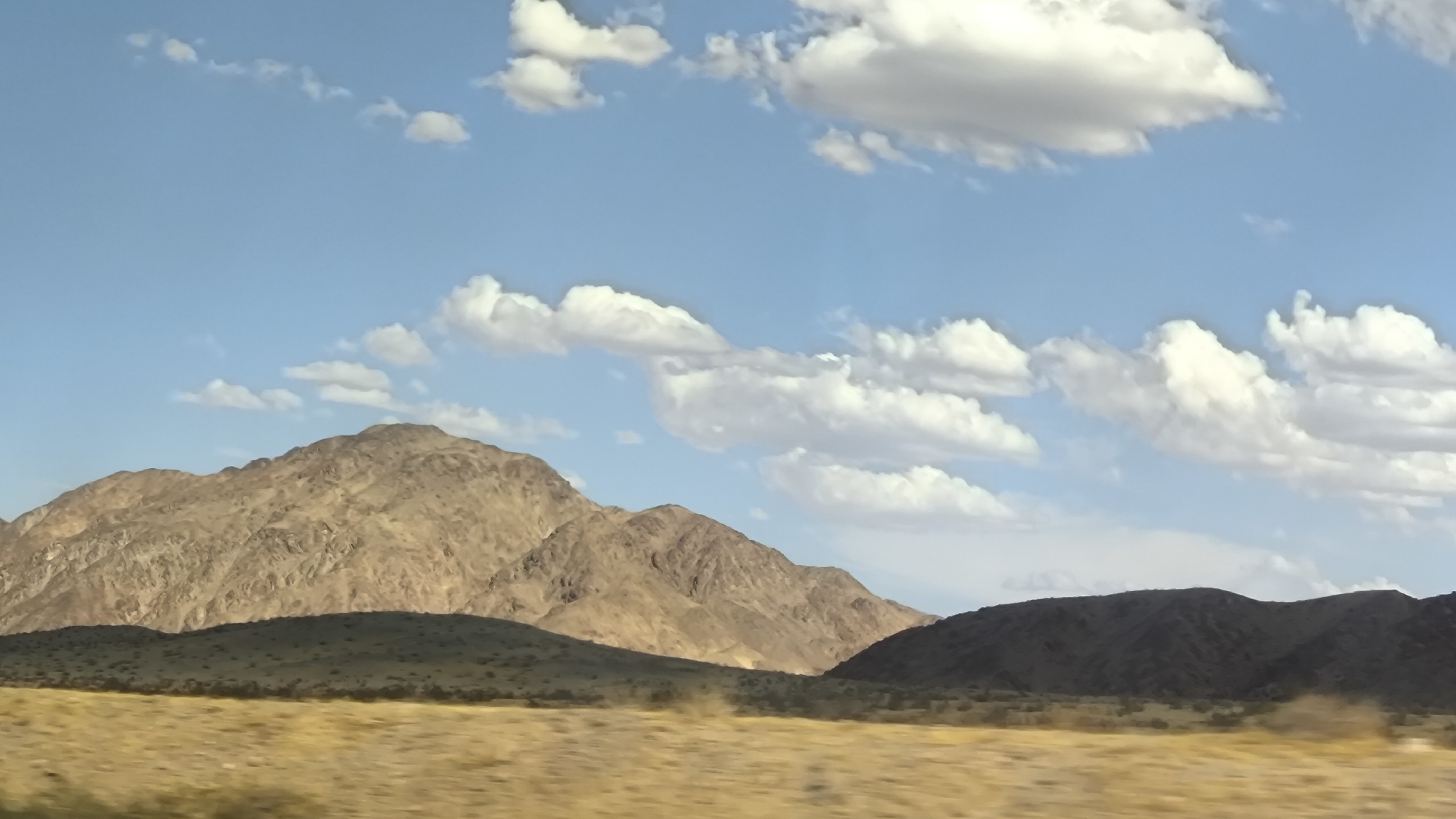 The image shows a desert landscape with mountains under a sky dotted with cumulus clouds. The foreground features dry, yellowish grass. In the middle ground, rolling hills with sparse vegetation are visible. The background is dominated by large, rugged mountains with textured surfaces, illuminated by sunlight. A series of smaller, darker hills are situated in front of the main mountain range. The sky is a clear blue with fluffy white and gray clouds scattered across it. The scene appears to be a natural outdoor environment. This image was captured in Afton, United States.