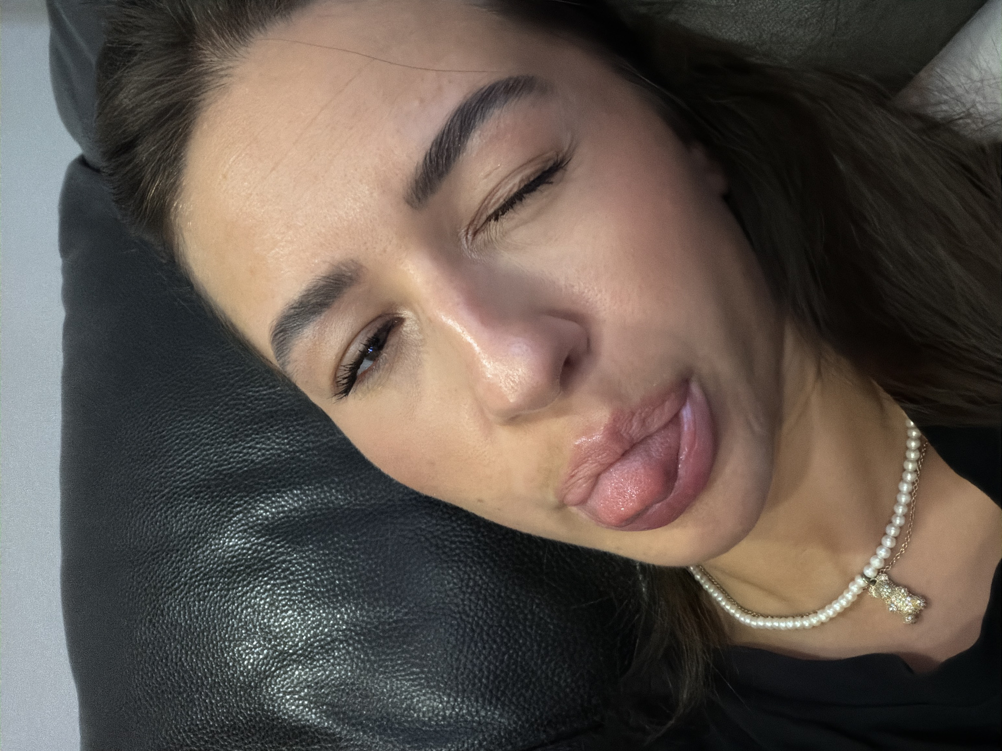 In Novi Iskar, Bulgaria, a woman with dark hair lies on a black leather surface, her face angled towards the camera. She is wearing a black garment and a pearl necklace with a teddy bear pendant. Her right eye is closed in a wink, while her left eye is open. Her tongue is extended, touching her lower lip. The lighting highlights the contours of her face and the texture of the leather.