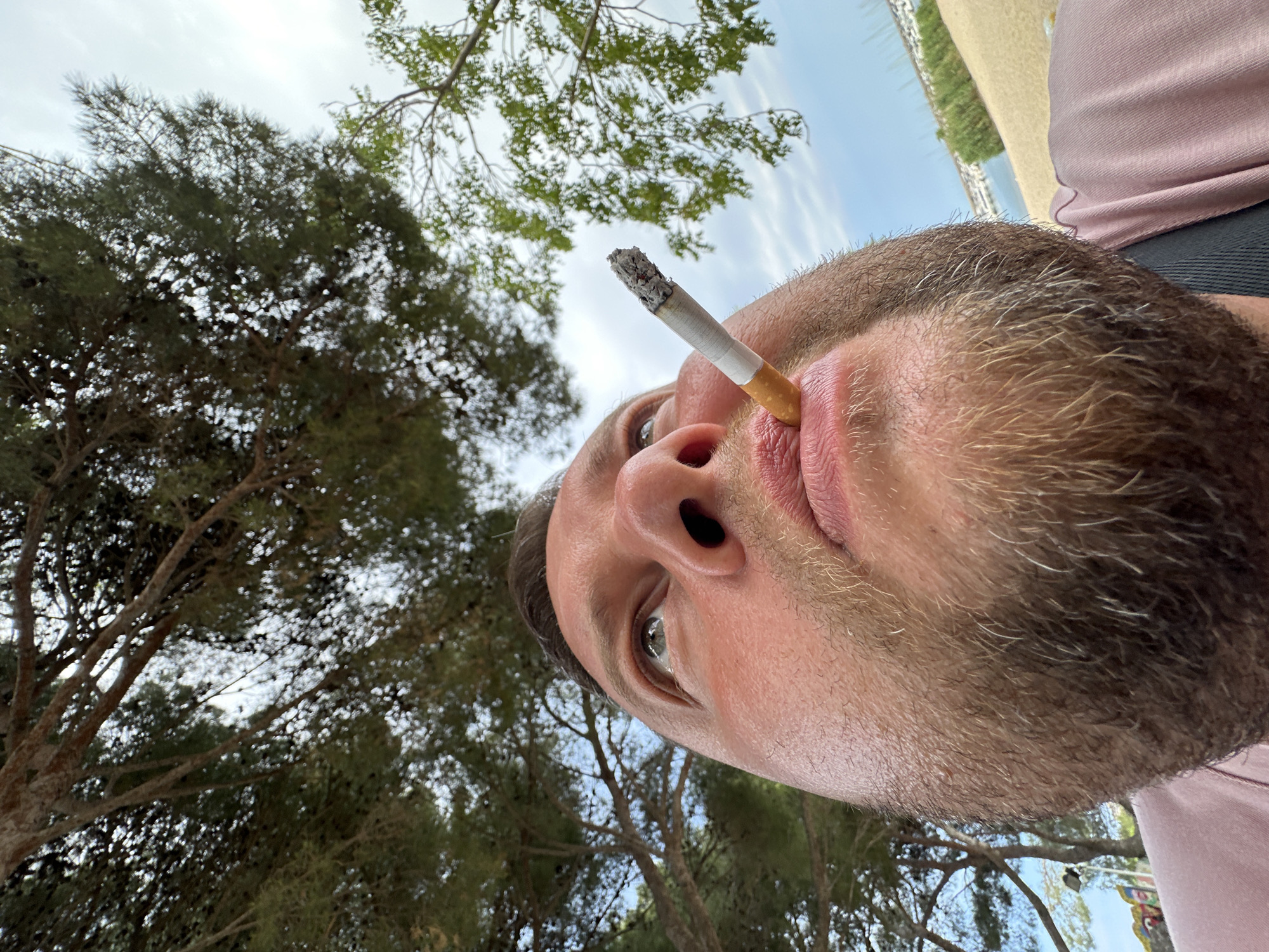 A low-angle, close-up, outdoor, waist-up shot captures a Caucasian man holding a lit cigarette in his mouth. The man is positioned in the foreground, with his face tilted upwards towards a canopy of pine trees. His skin appears flushed, and he has a short, stubbly beard. The cigarette, with ash at its tip, is held between his lips. In the background, out of focus, is a sandy beach bordering a body of water, likely the Mediterranean Sea, with some boats and structures visible in the distance. The sky is overcast. This scene is likely located in Sant Feliu de Guíxols, Spain, a coastal town known for its beaches and pine forests.