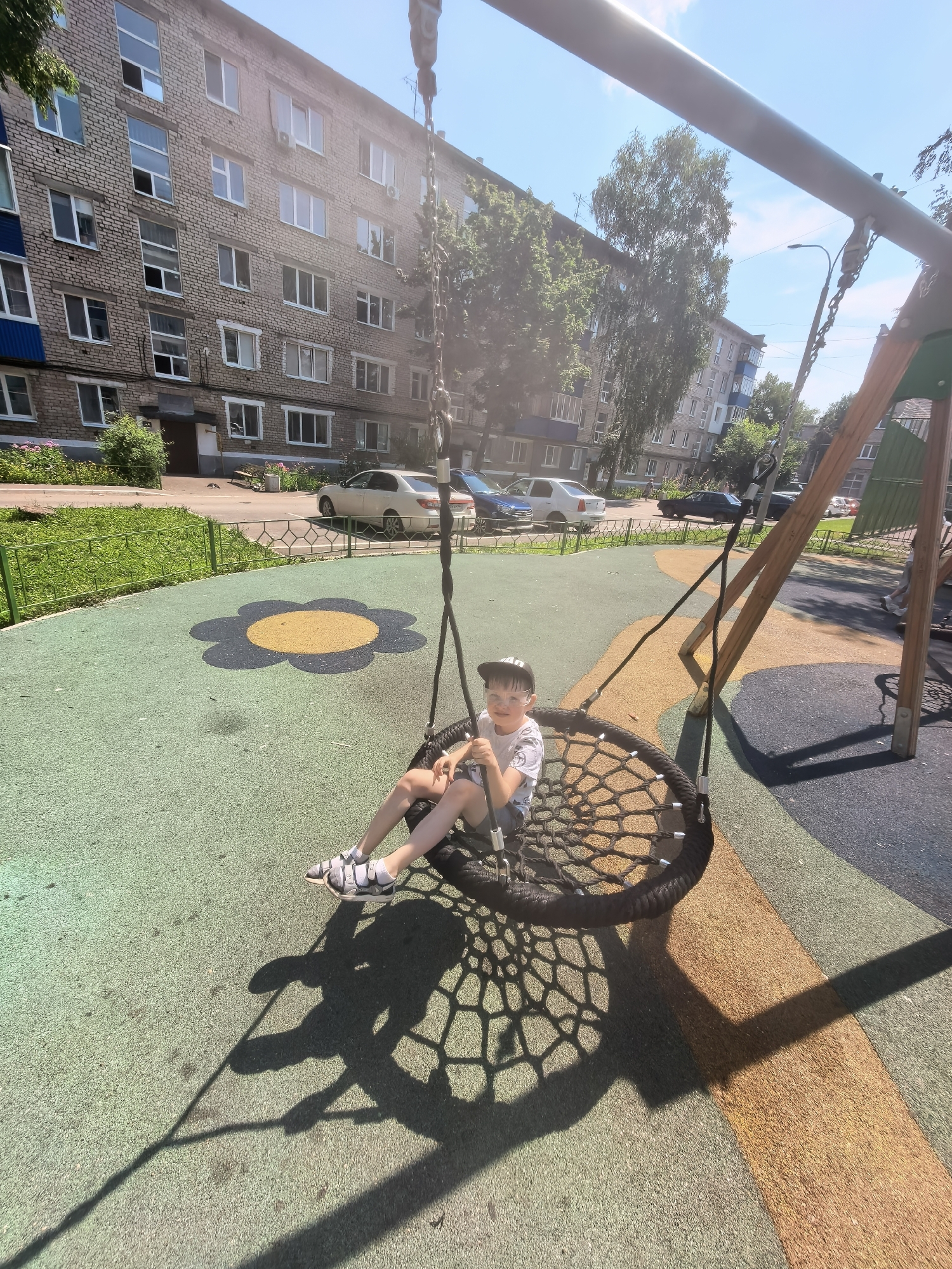 A boy with a hat and glasses is sitting on a large, round swing in a playground. He is wearing a t-shirt and shorts. The playground has a colorful rubberized surface with a flower-shaped pattern. In the background, there is a multi-story brick apartment building with many windows, characteristic of urban residential areas in Sterlitamak, Russia. Trees and parked cars are also visible. The sun is shining, casting shadows on the playground.