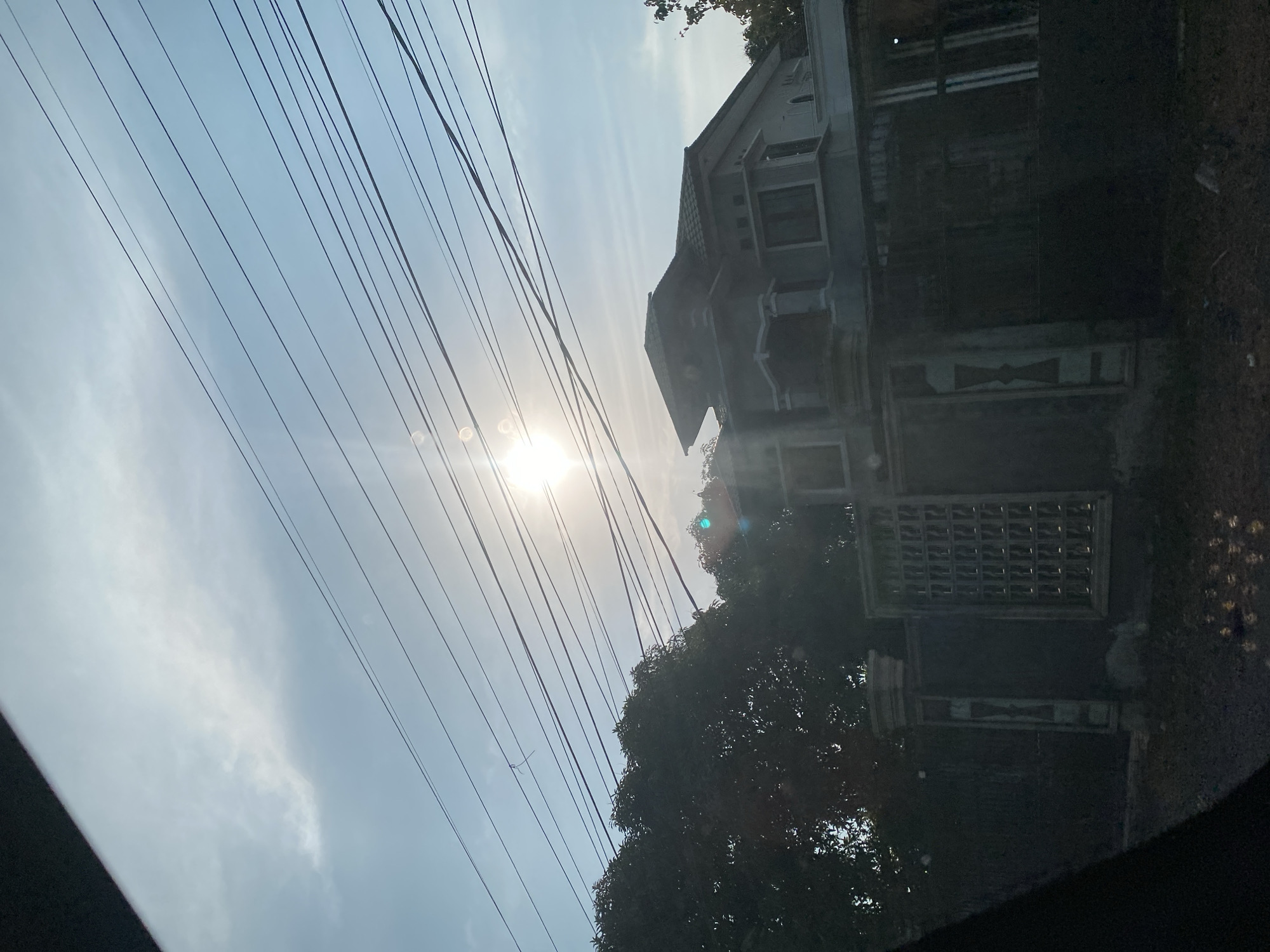In the city of Jati Baru, Indonesia, an outdoor scene is captured during daylight hours, bathed in the bright glare of the sun. The sky above is a light blue, streaked with wispy white clouds and dominated by power lines that crisscross the frame horizontally. A large, leafy tree, dark in silhouette, occupies the left side of the image, providing a stark contrast to the luminous sky. To the right of the tree, a modern-looking house with a multi-tiered roof and visible windows is partially obscured by the tree and the power lines. A decorative fence with geometric patterns and pillars runs in front of the house. The foreground is dark and out of focus, with some debris visible on the ground. The overall impression is one of a residential area under a clear, sunny sky.