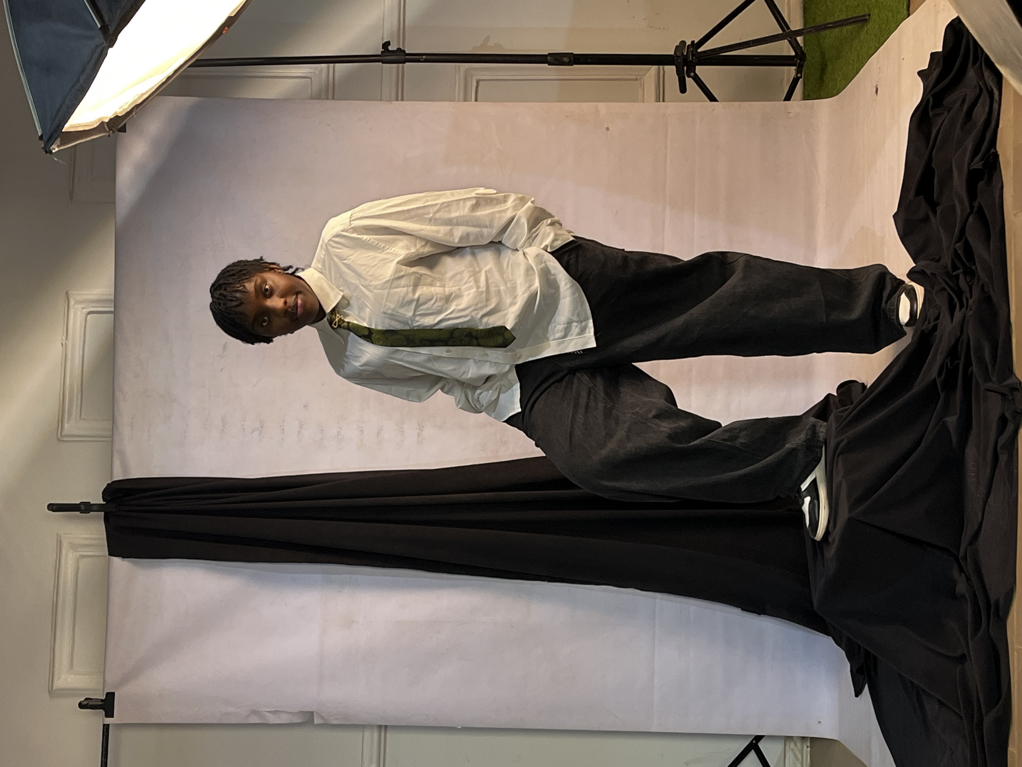 A photograph taken in what appears to be a studio setting, showing a person posing against a white backdrop. The individual is wearing a white button-down shirt with a tie and dark-colored pants. The person is looking at the camera. The backdrop is partially covered by draped black cloth. A lighting setup, including a softbox, is visible in the upper left corner, and a tripod is partially visible in the upper right corner. The location in Barakin Akawo, Nigeria, cannot be confirmed from the image alone.