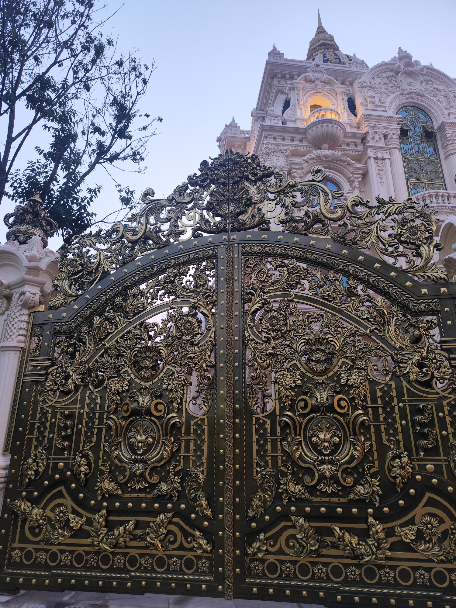 The image shows an ornate, gold and dark metal gate in front of an equally ornate building in Da Lat, Vietnam. The gate is highly detailed, with swirling floral and leaf designs, as well as decorative circular emblems. The building behind the gate is multi-storied and constructed in a pale pink color. It is elaborately adorned with sculpted elements, including columns, arched windows, and a prominent dome at the top. A tree with bare branches is visible to the left of the gate. The sky appears to be a pale, overcast color, suggesting either early morning or late afternoon. The photo is taken from a low angle, emphasizing the height and grandeur of both the gate and the building.